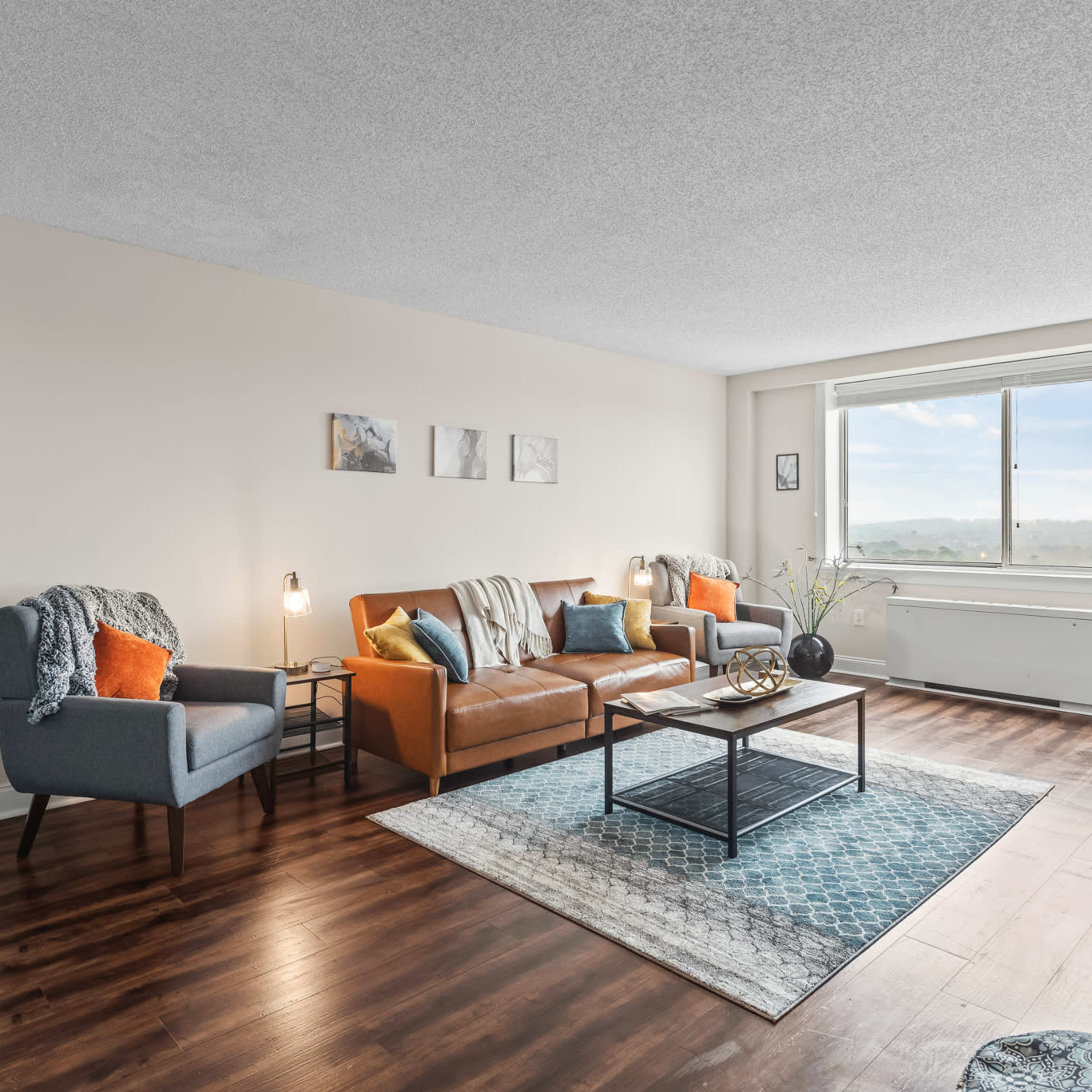 A modern living room features a brown leather couch, an armchair, and a coffee table, with a large window providing ample natural light.