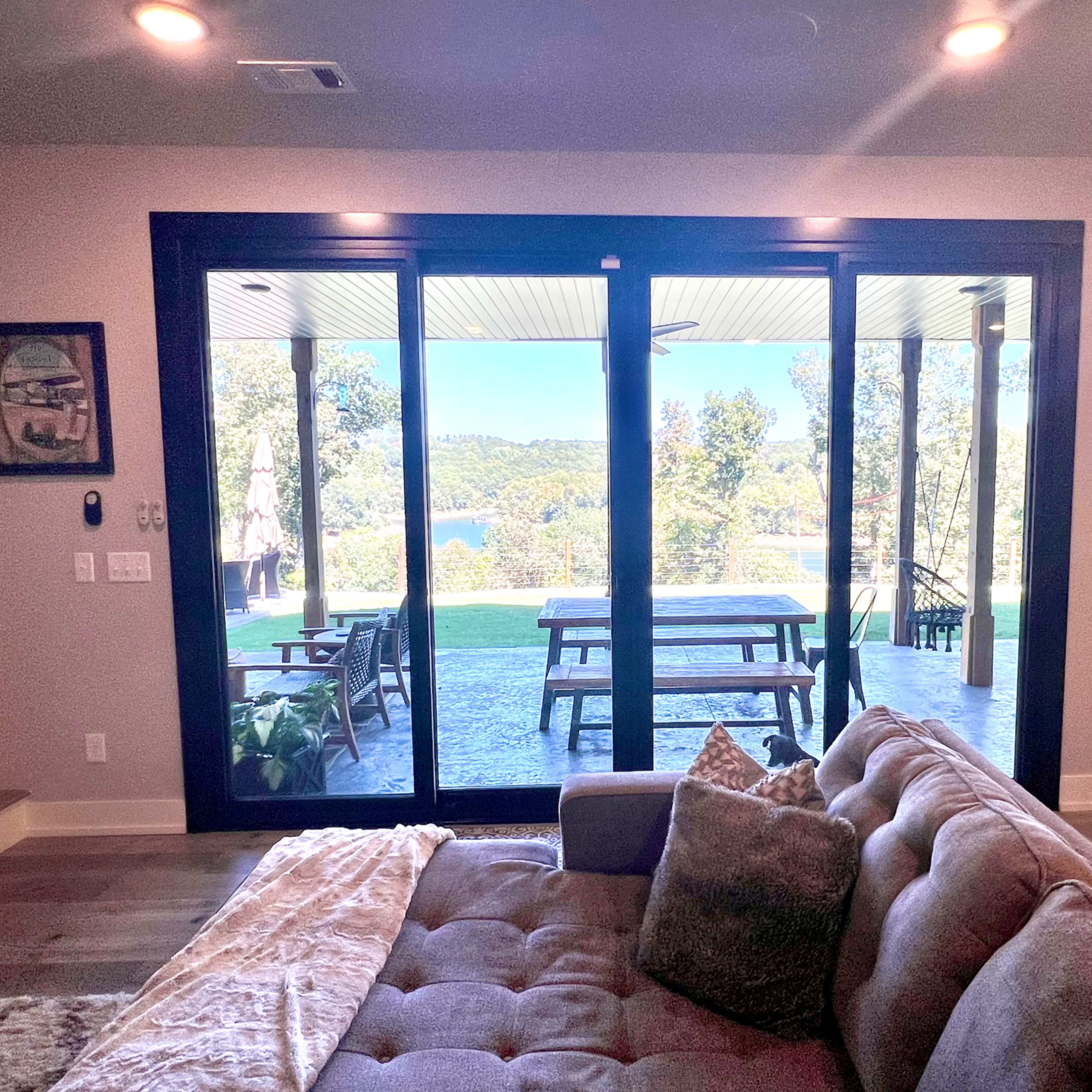 A view of a living room with a couch, leading to large sliding glass doors that open to a patio and a scenic landscape with trees and a body of water.