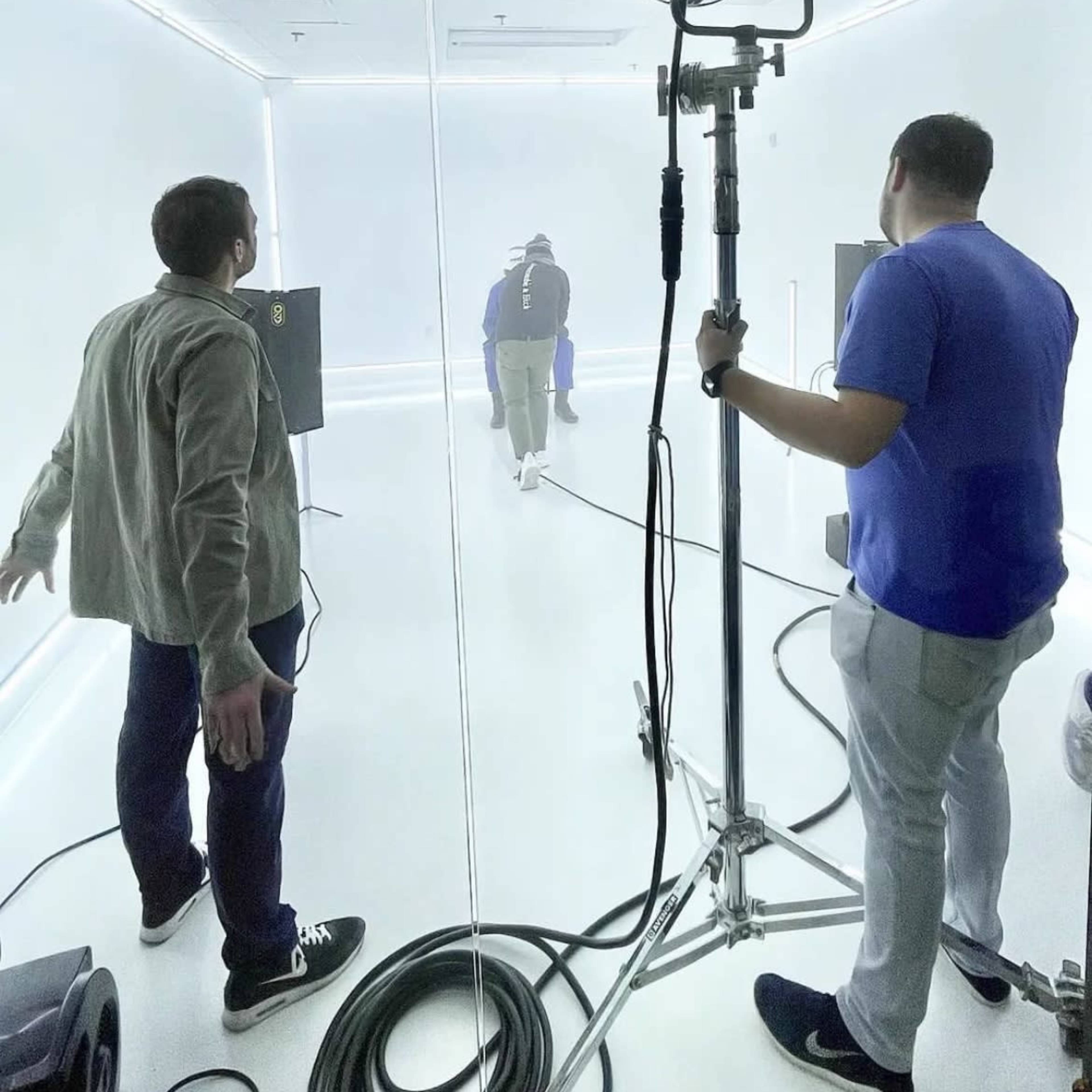 Two crew members are positioned in a brightly lit studio, while a third person walks away into a light-filled area.