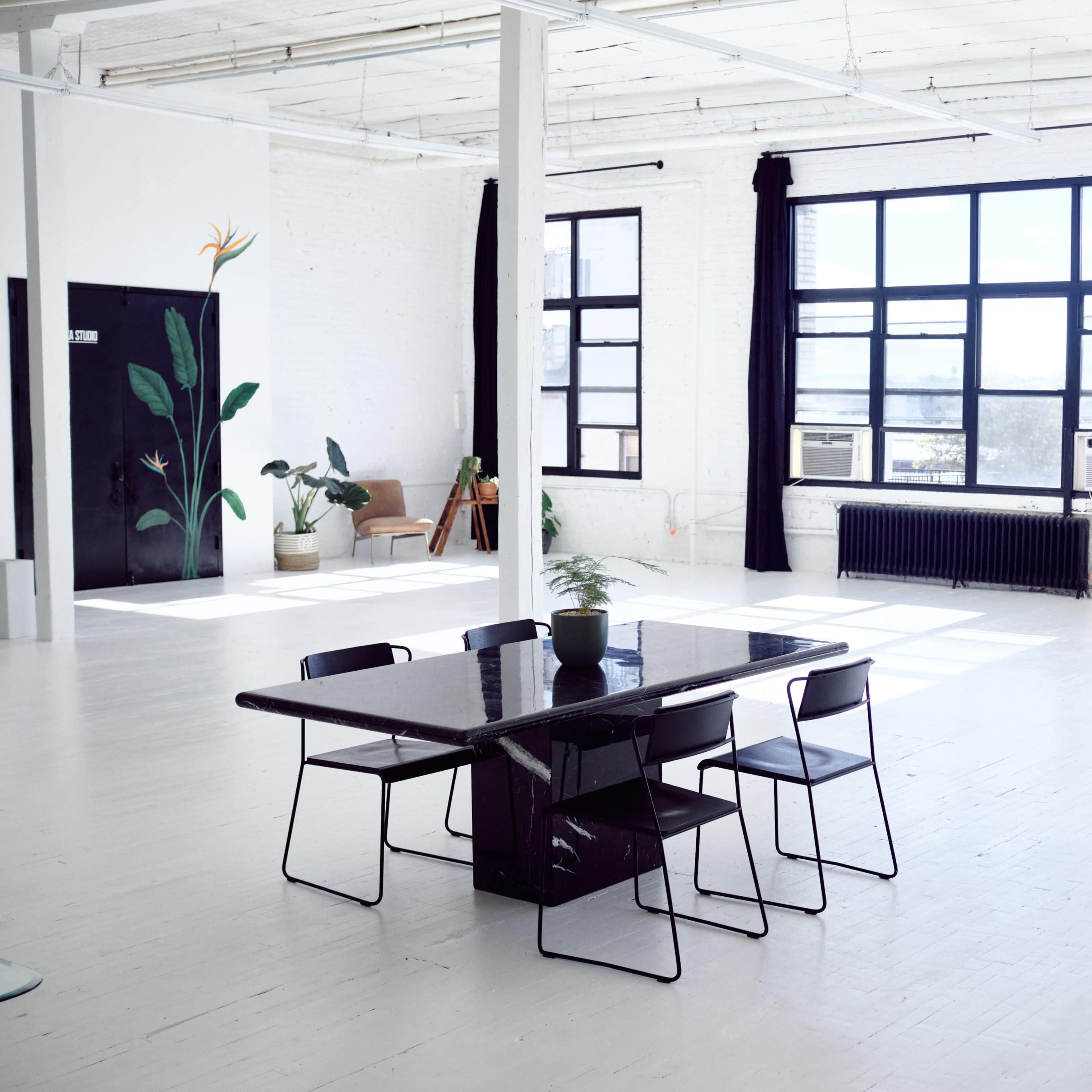 A spacious, well-lit room with large windows, featuring a black table with chairs and a plant.