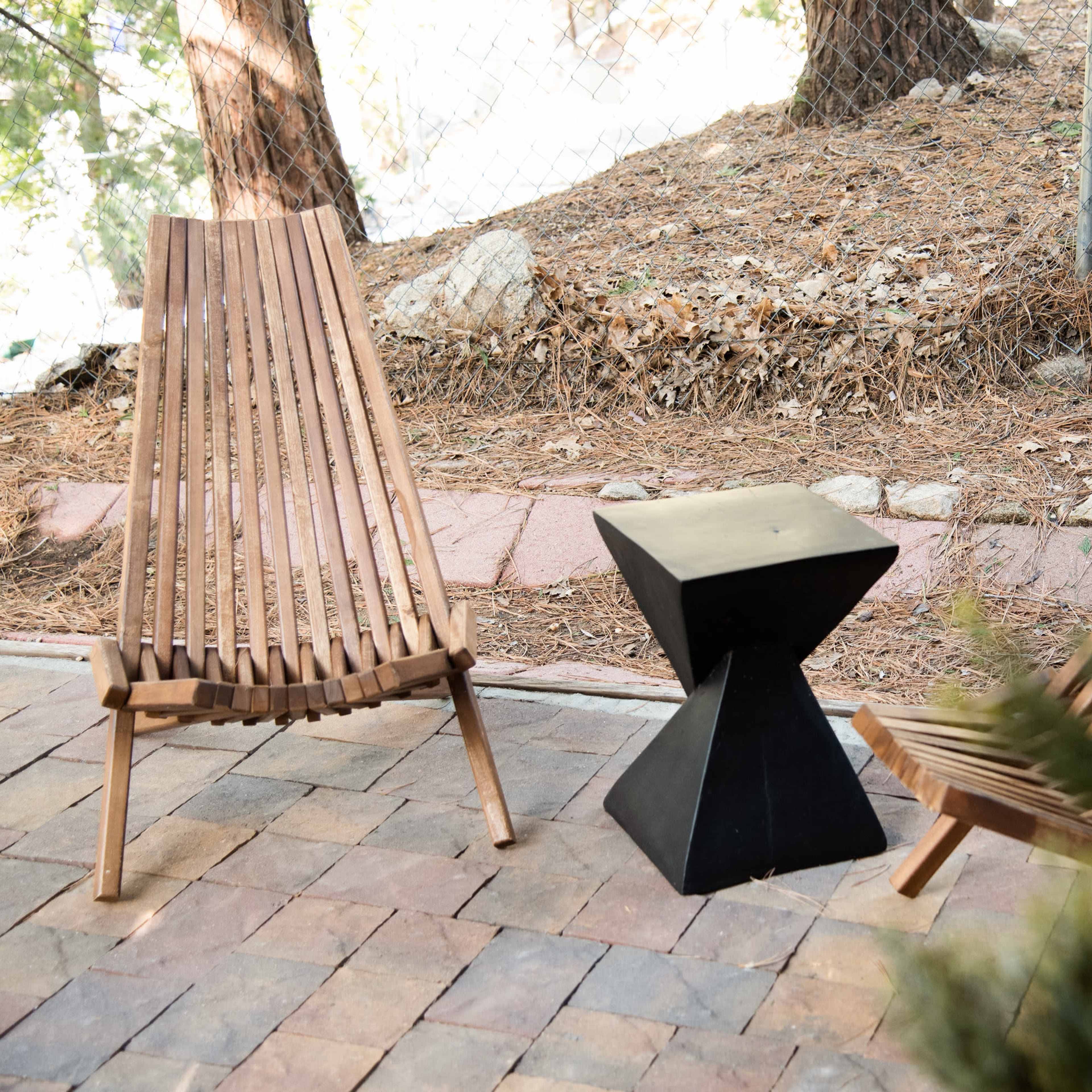 Two wooden lounge chairs are positioned beside a black geometric side table on a stone-paved area near a wooded backdrop.