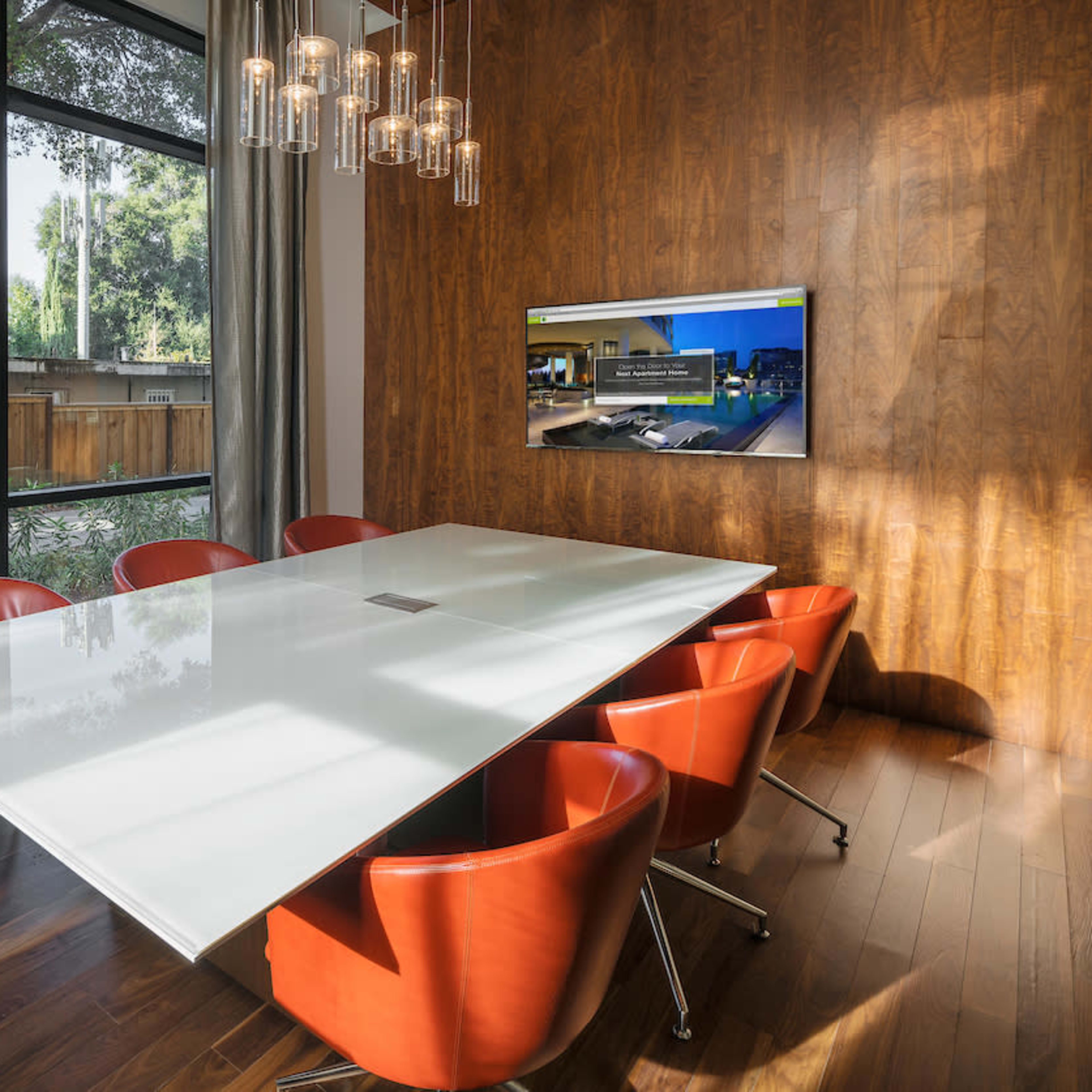 A modern conference room features a long white table surrounded by red chairs, with large windows offering a view of the outdoors and a television mounted on a wooden wall.