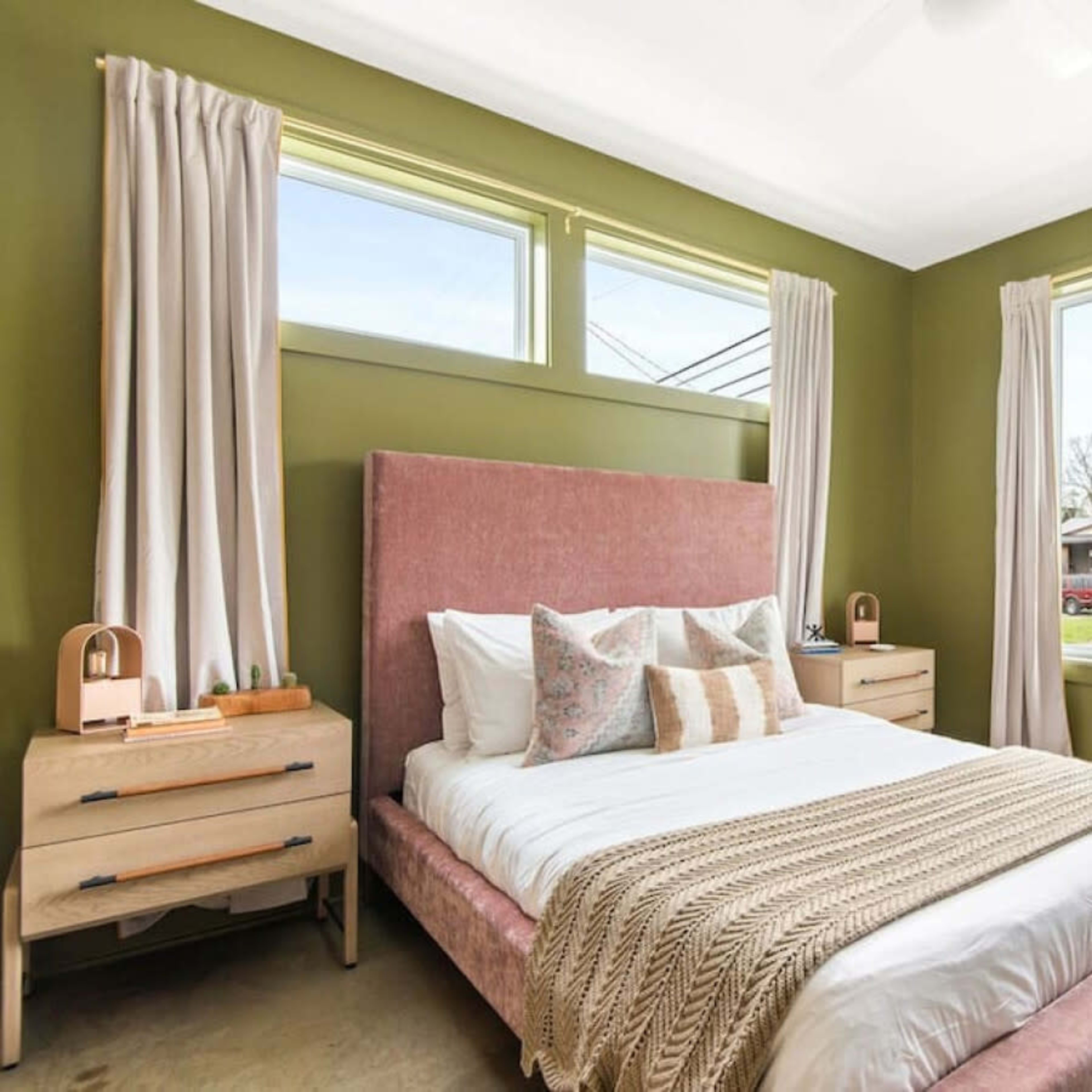 A bedroom with a pink upholstered bed, light-colored bedding, wooden nightstands, and large windows that provide natural light.