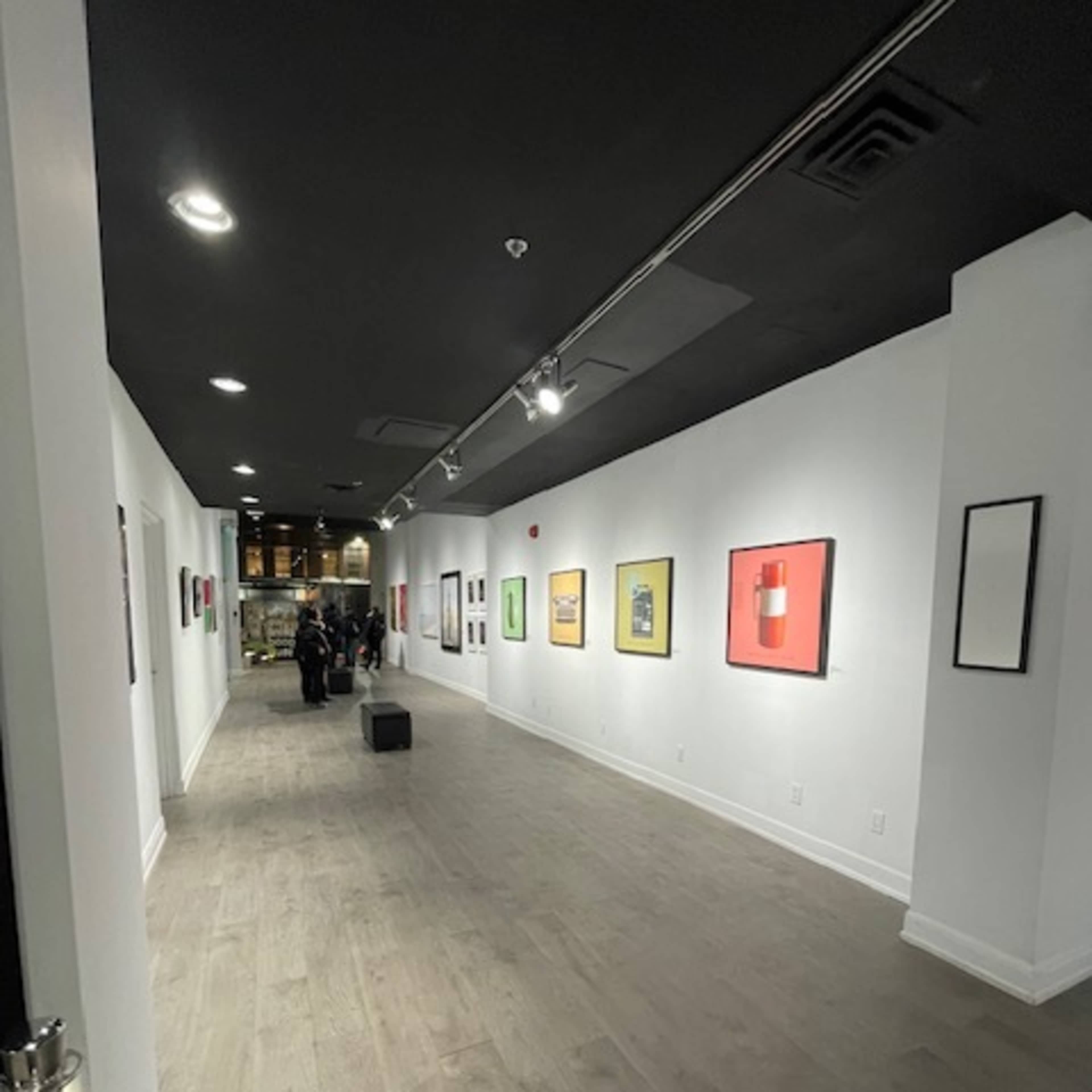 A long, well-lit art gallery hallway with framed artworks displayed on the walls and a group of people in the background.