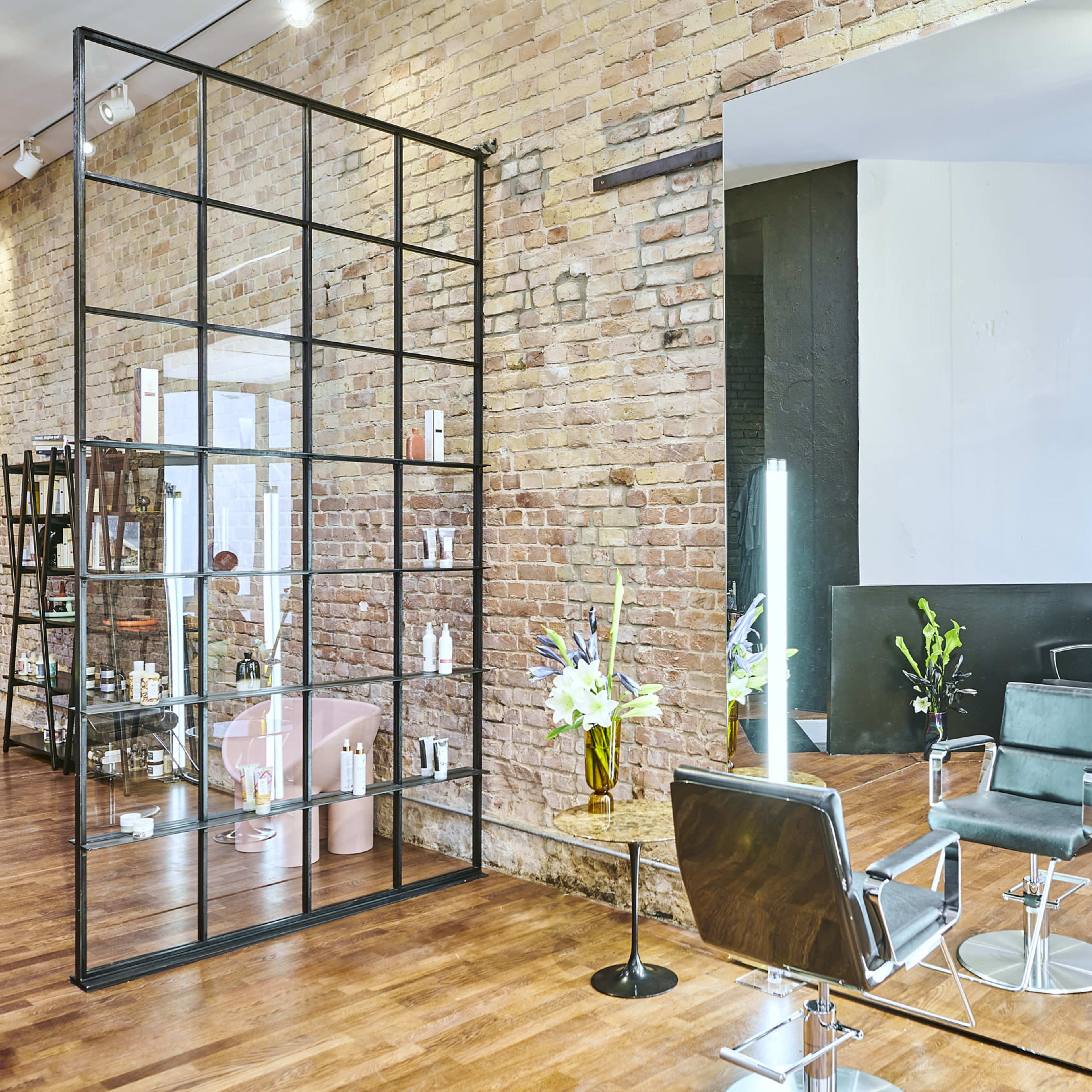 A modern salon interior with exposed brick walls, glass partitions, and salon chairs positioned near a large mirror.