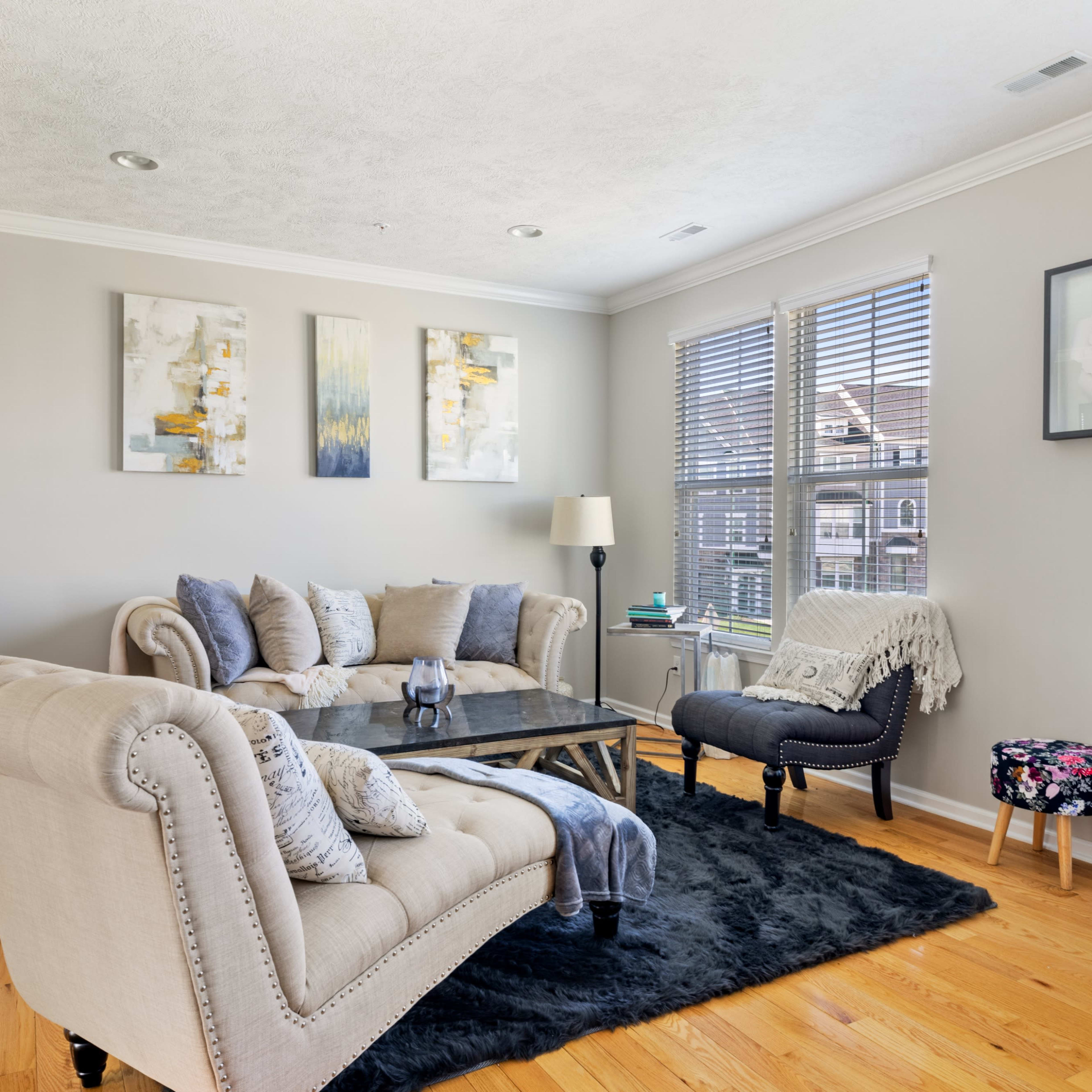 A living room features a beige sofa and armchair, a glass coffee table on a black rug, and wall art above, all illuminated by natural light from windows.
