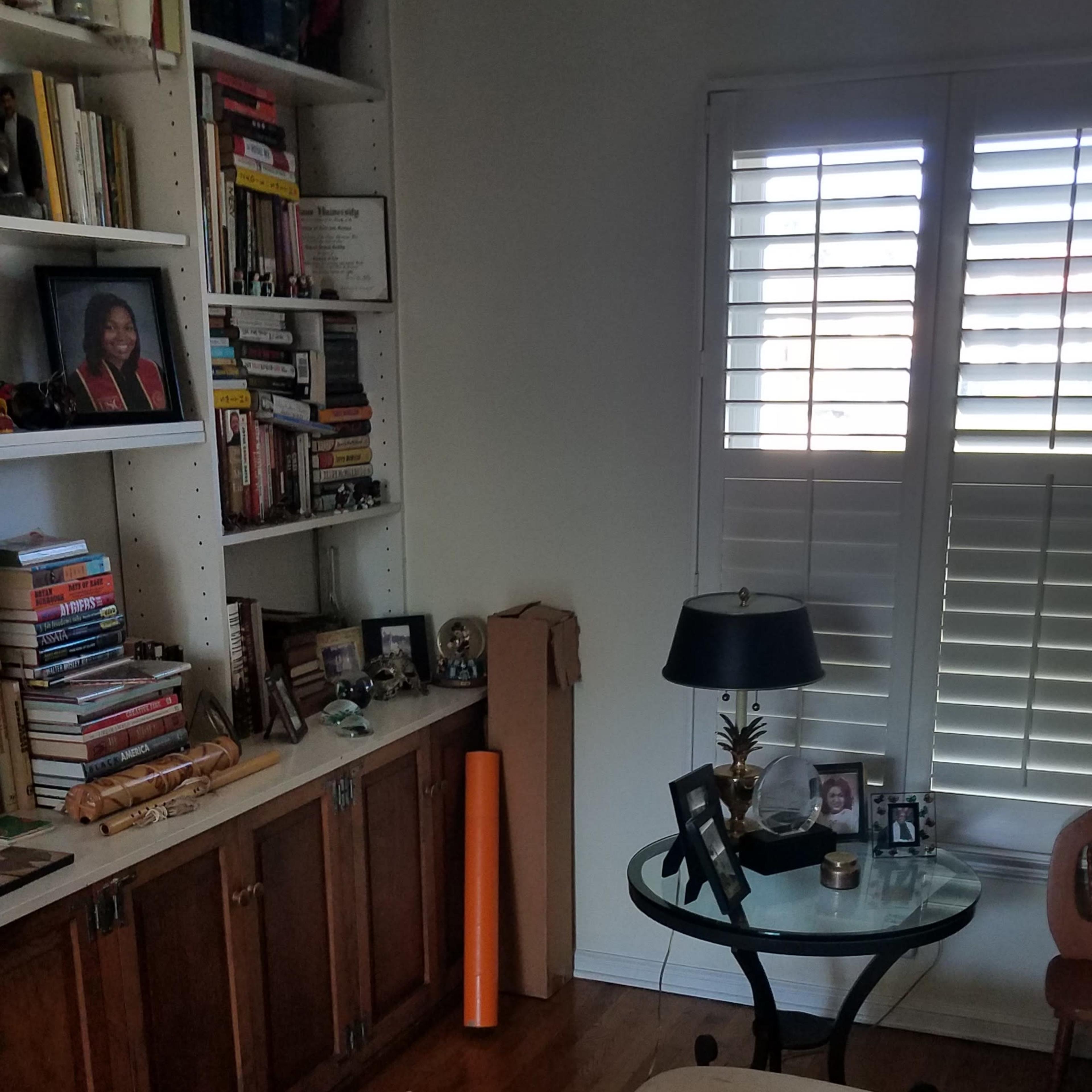 A cozy room features a bookshelf filled with books, a coffee table with a decorative tray and flowers, and a window with plantation shutters.