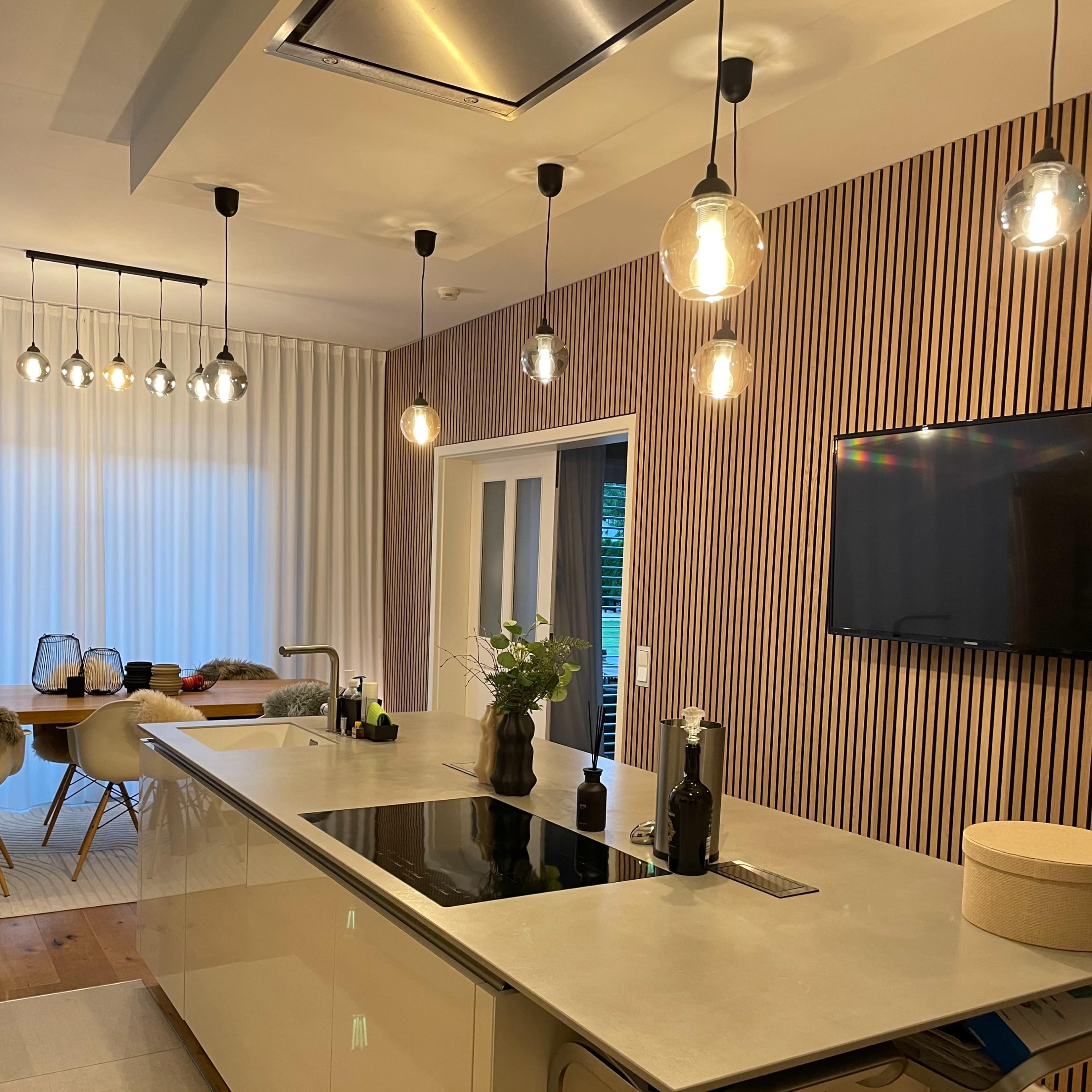 A modern kitchen with a central island surrounded by pendant lights, featuring a sleek countertop and a wall-mounted television.