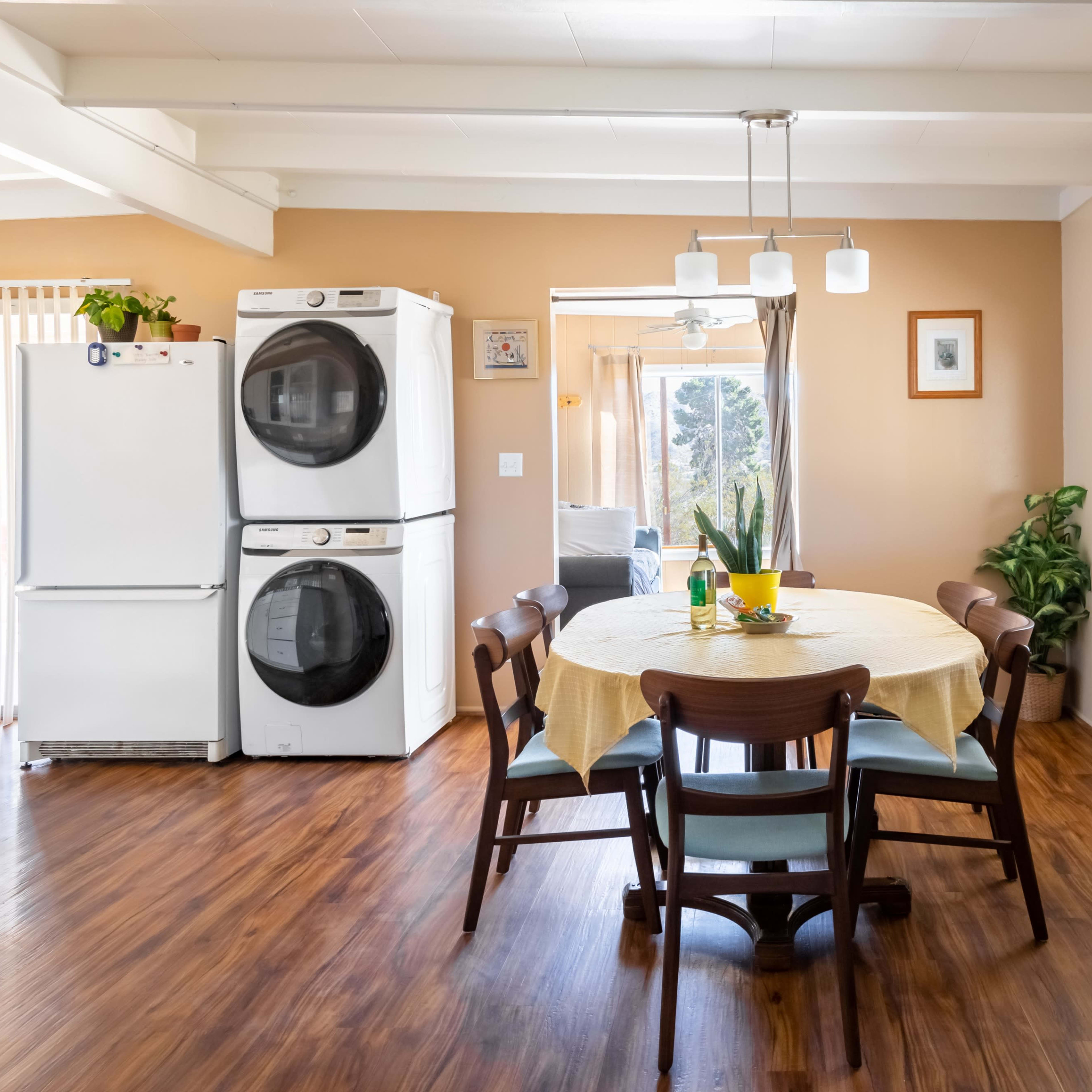 A dining area features a round table surrounded by chairs, with white appliances and large windows providing natural light.