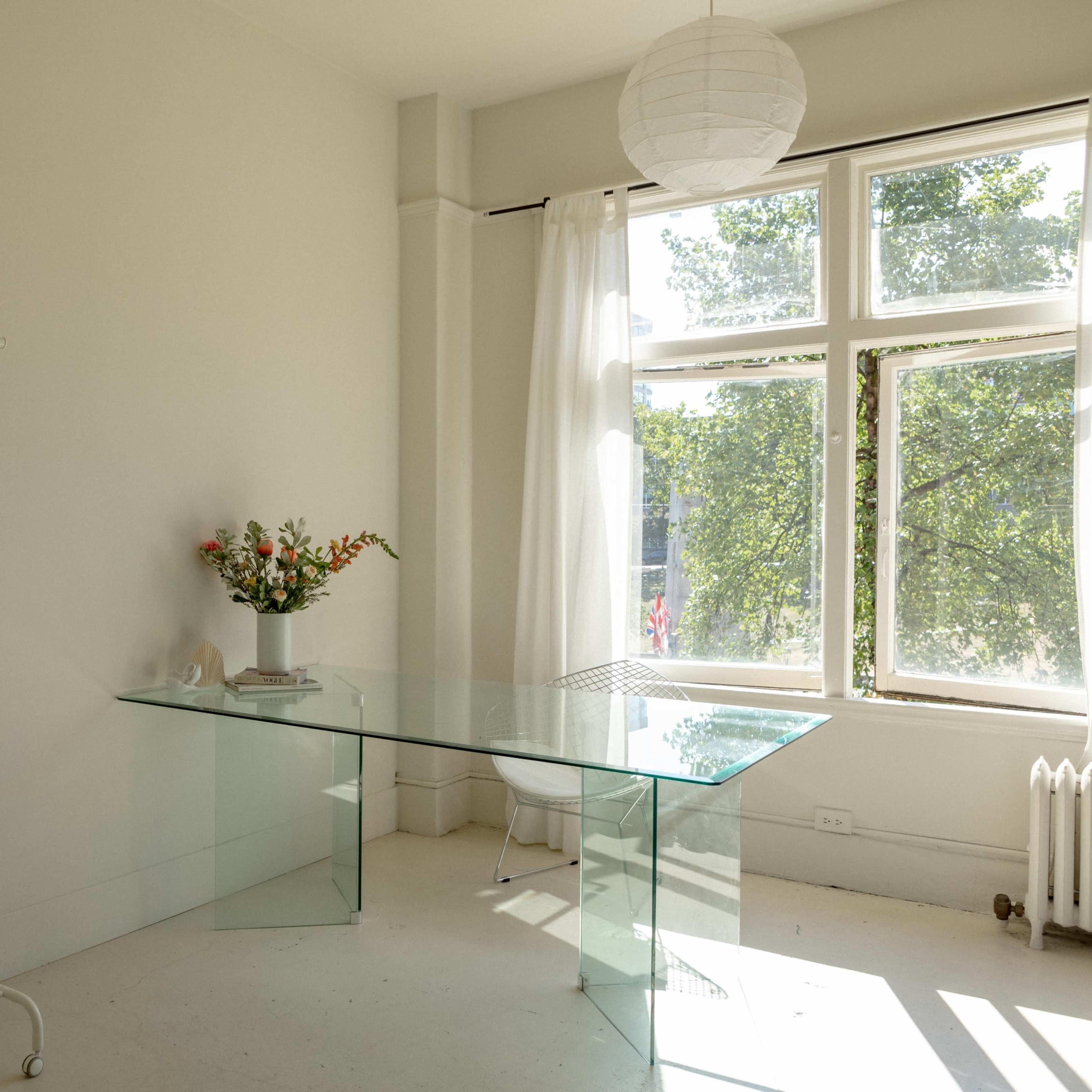 A minimalist room with a glass table, a clothing rack with hangers, and a vase of flowers on the table, illuminated by natural light from a large window.