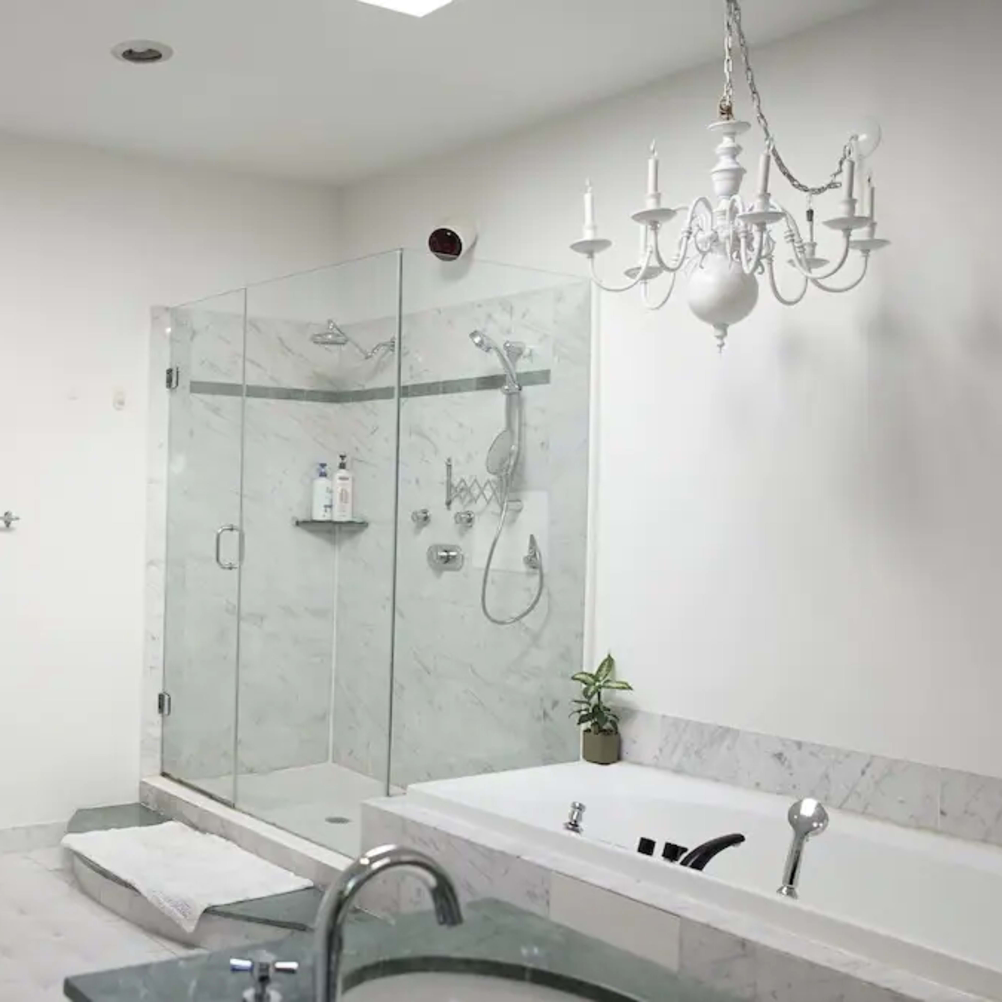 The bathroom features a glass shower, a large bathtub, and a chandelier, with an open doorway leading to another room.
