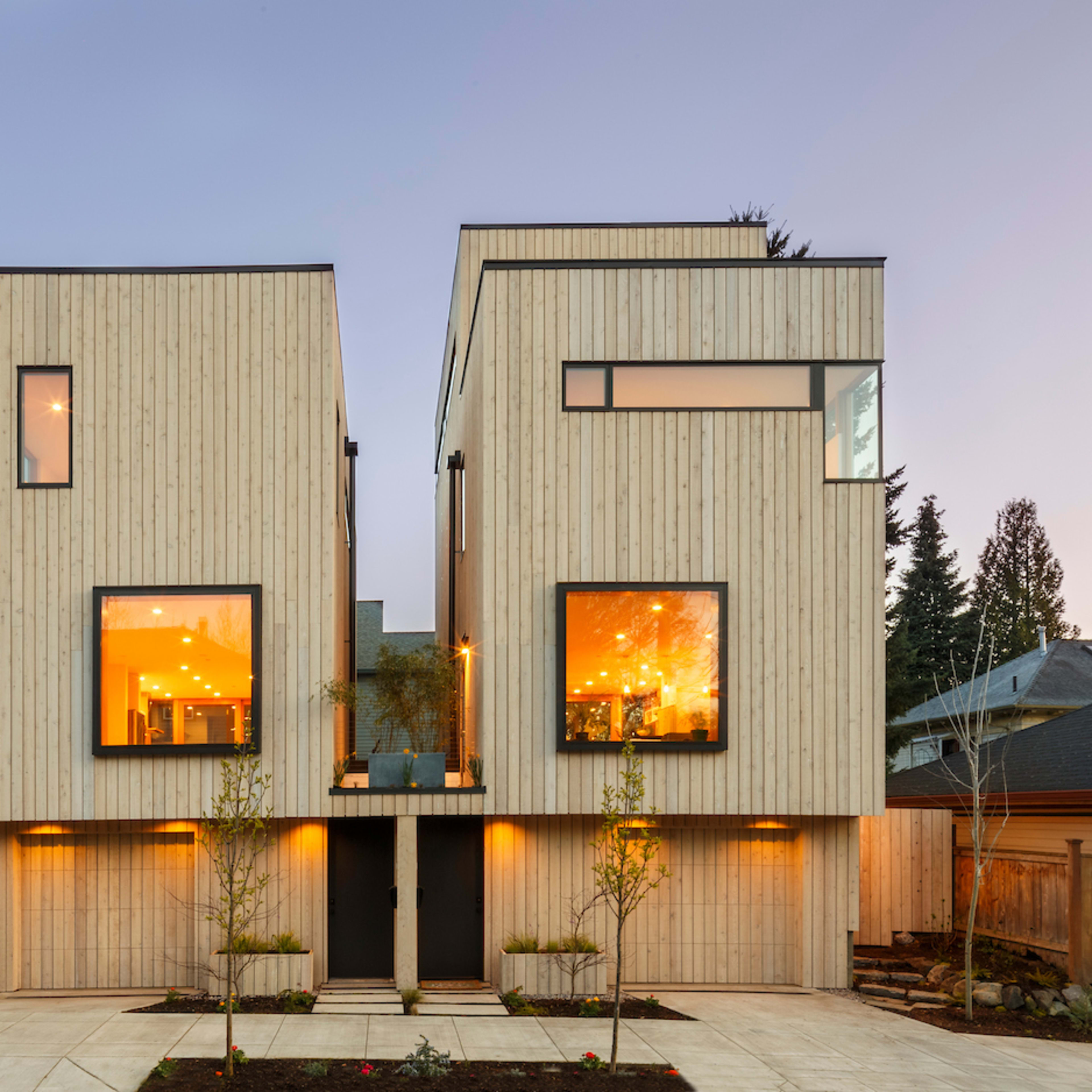 A modern, two-story wooden house with large windows, illuminated interiors, and a symmetrical facade.