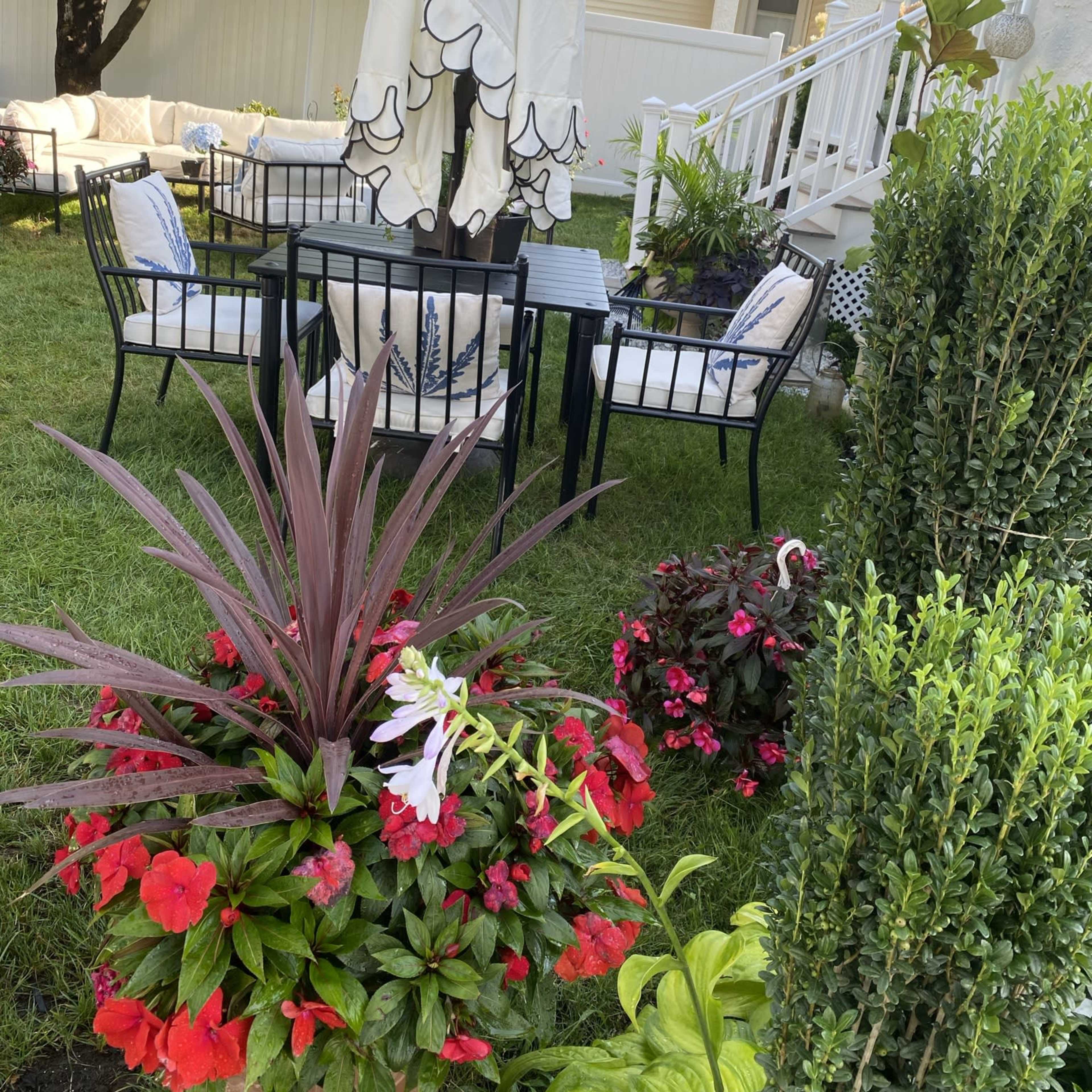 A backyard patio area with a table and chairs beneath a large umbrella, surrounded by various flowering plants.