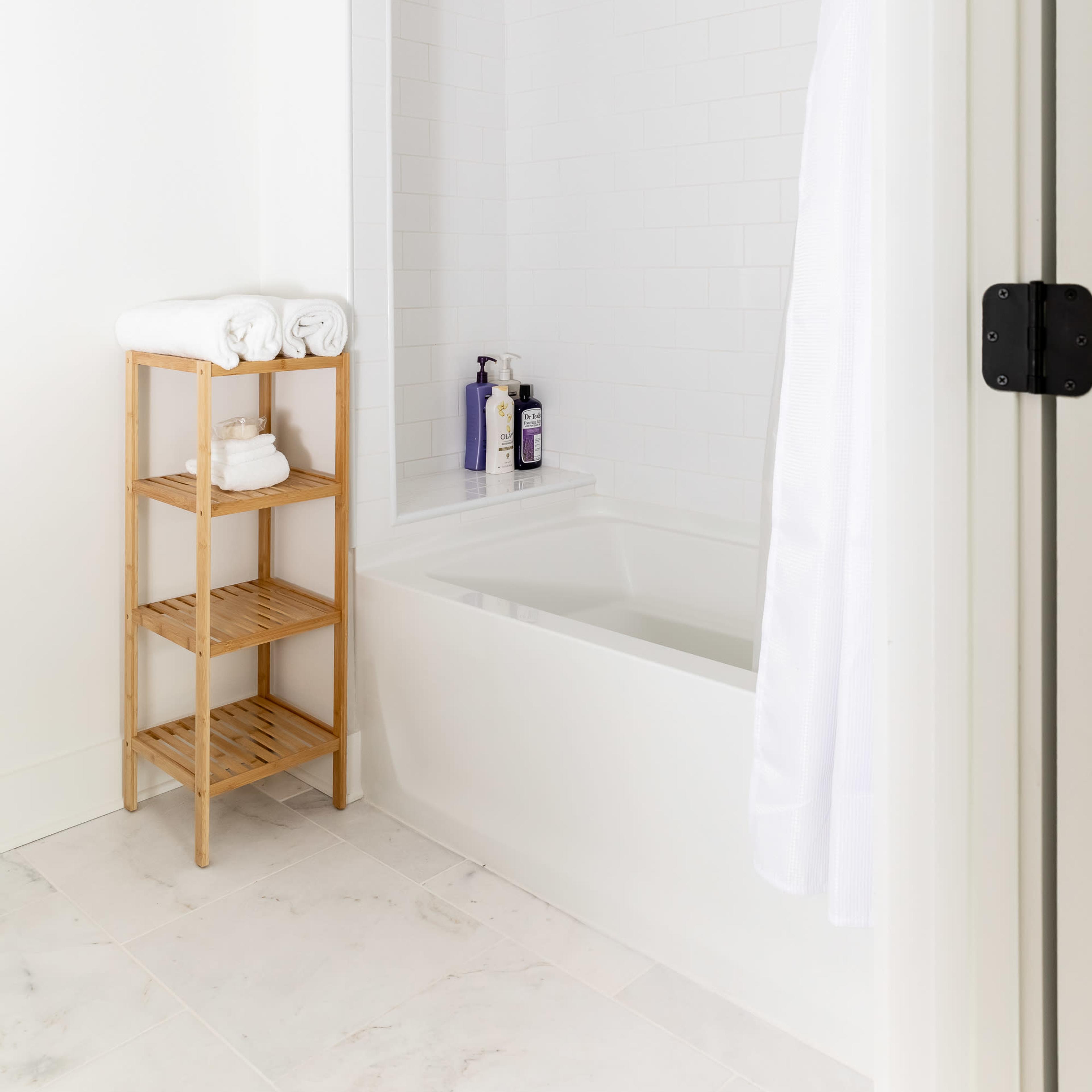 A clean bathroom featuring a bathtub with a shower curtain, a bamboo shelving unit with neatly stacked towels, and several bottles of toiletries.