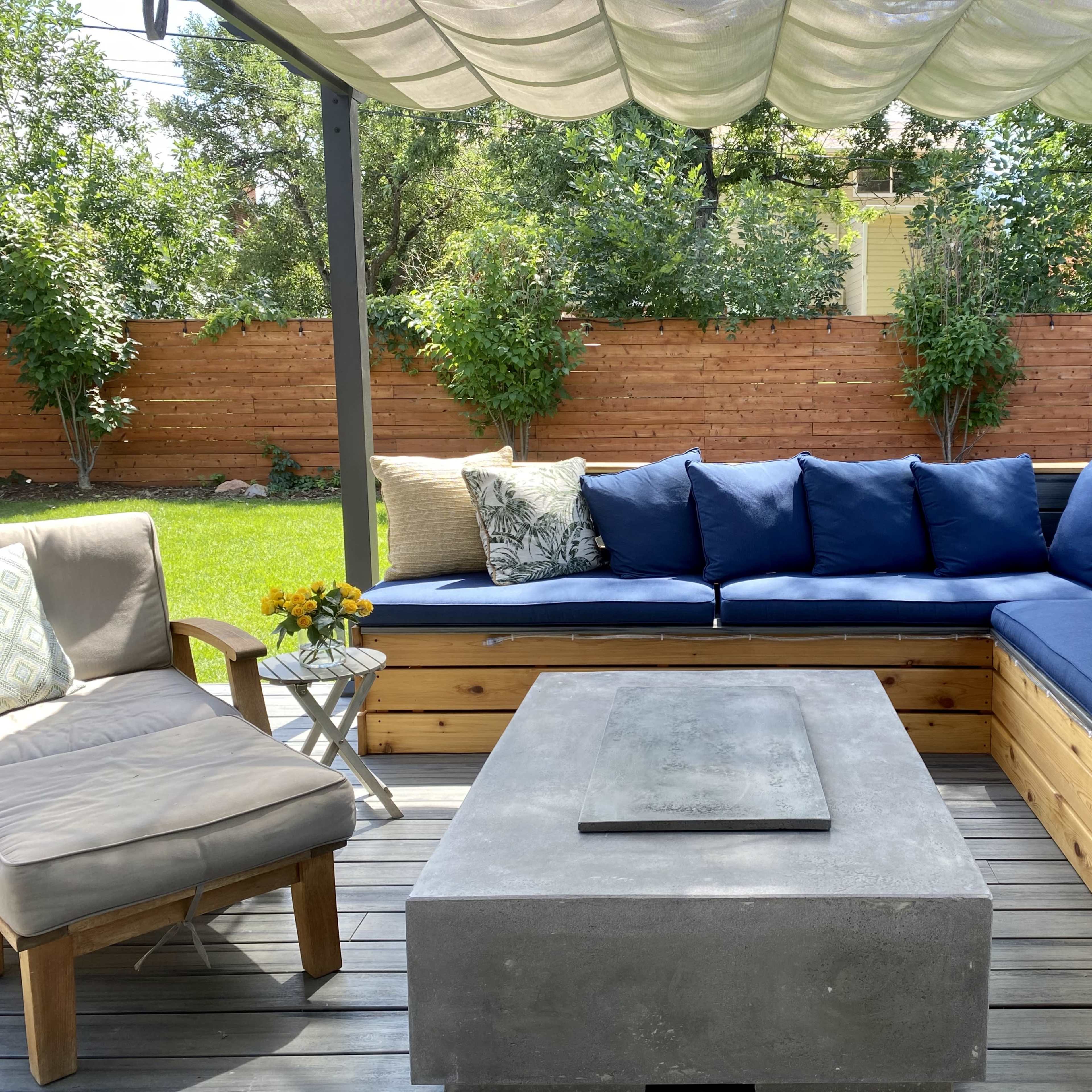 A outdoor seating area features a large sectional couch with blue cushions, a concrete coffee table, and a small side table, all under a fabric canopy.