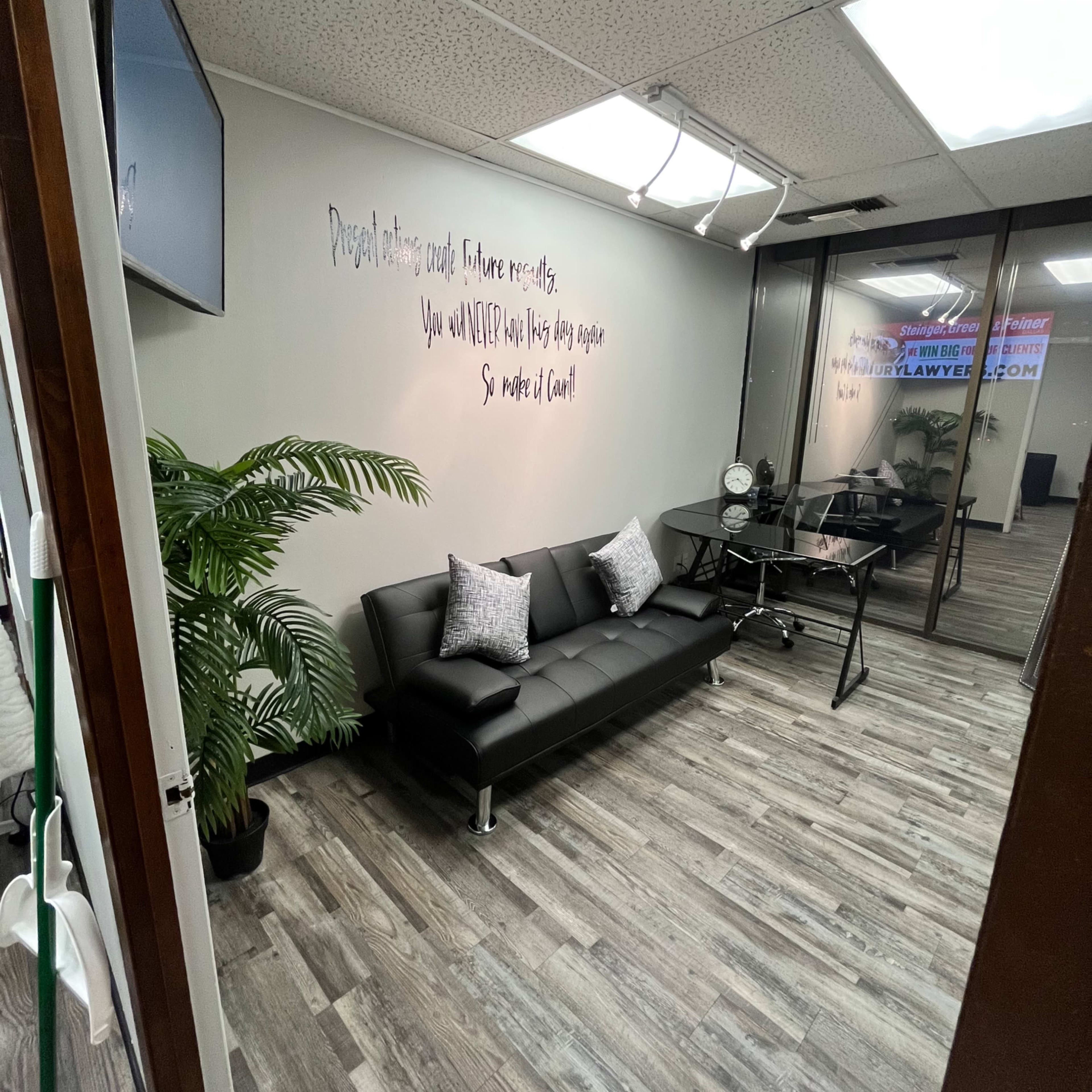 A modern office waiting area with a black sofa and a desk, featuring a large window and a decorative plant.