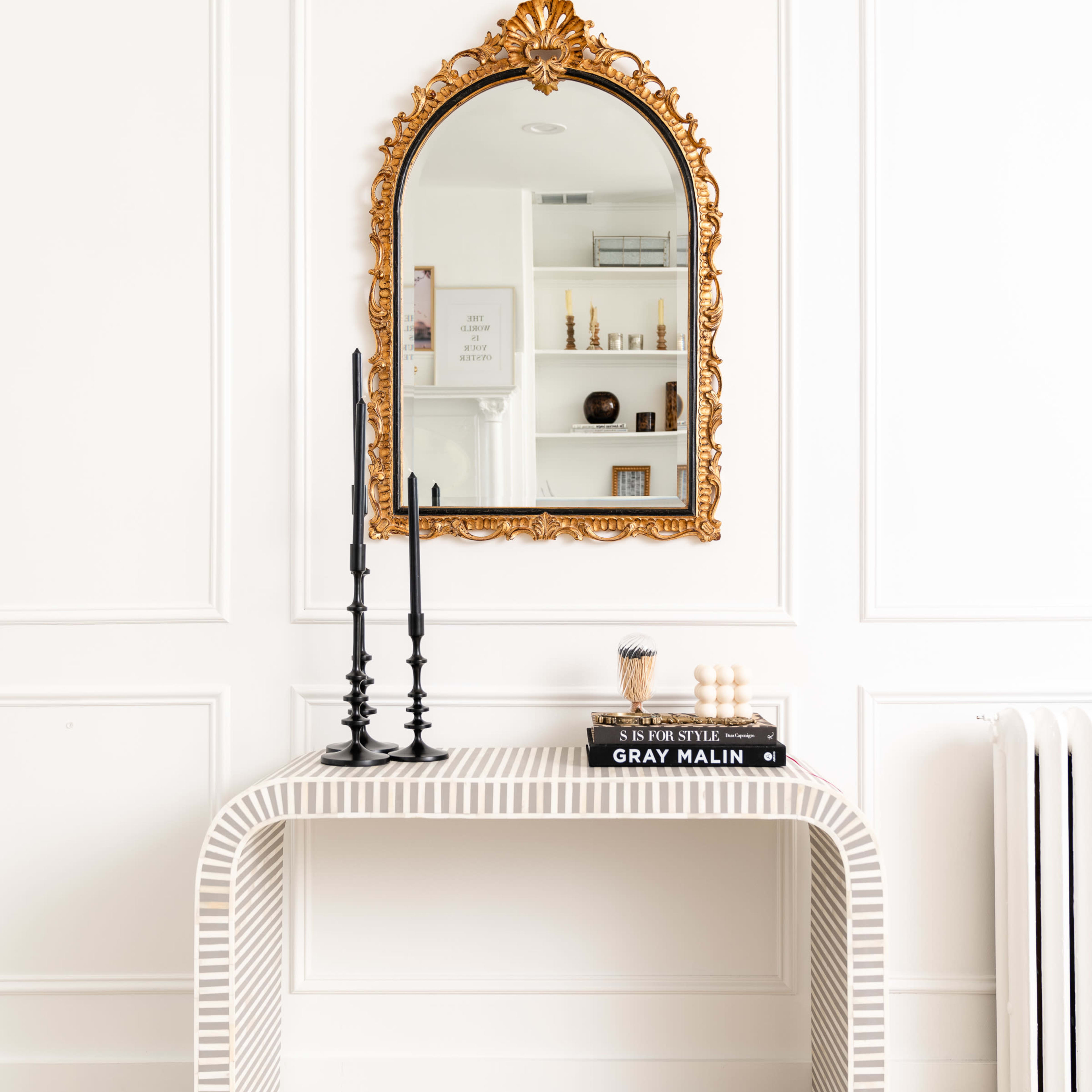 A striped console table with a gold-framed mirror and black candlesticks is positioned against a white wall.