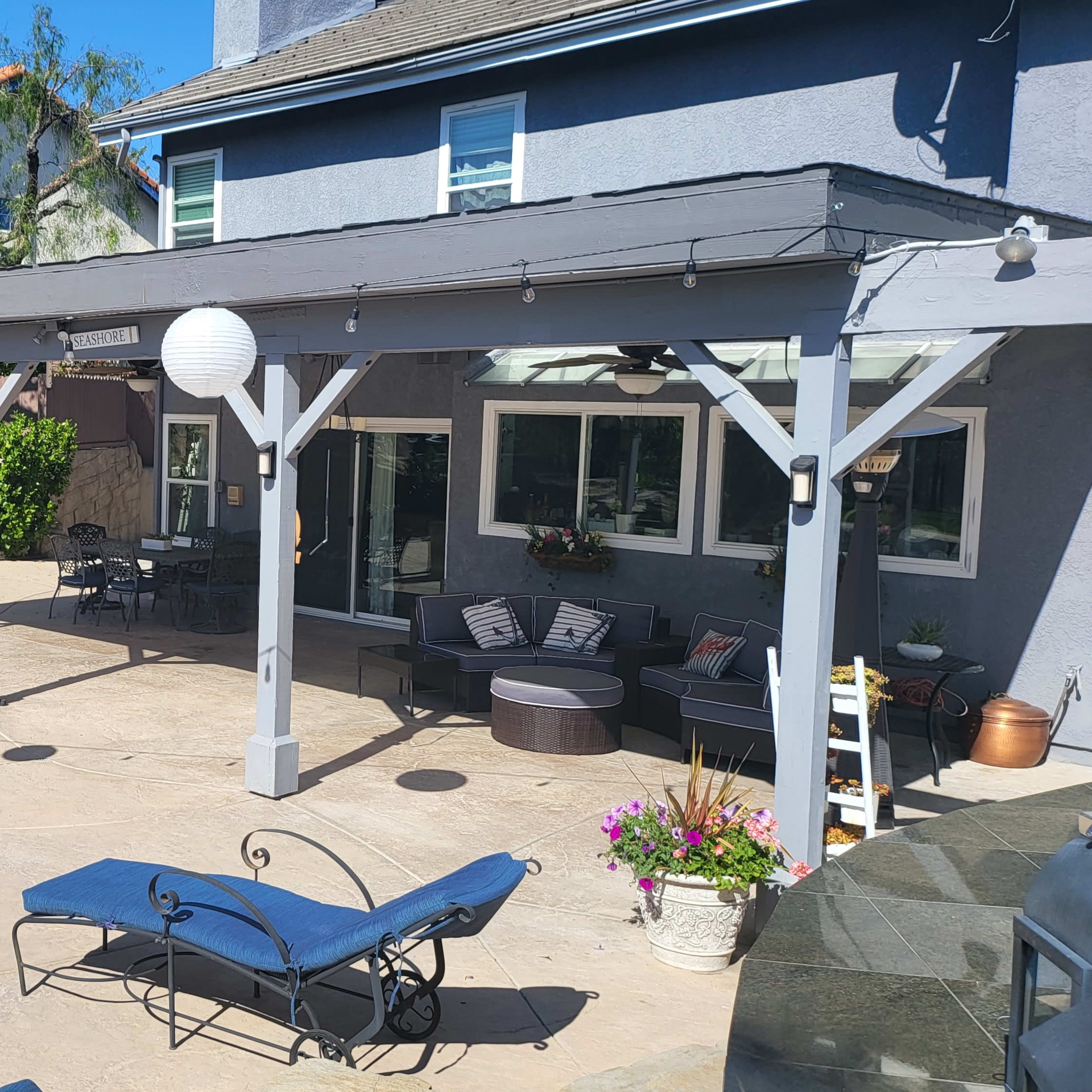 A spacious outdoor patio area with lounge chairs, potted plants, and decorative lanterns under a shaded structure adjacent to a pool and landscaped yard.