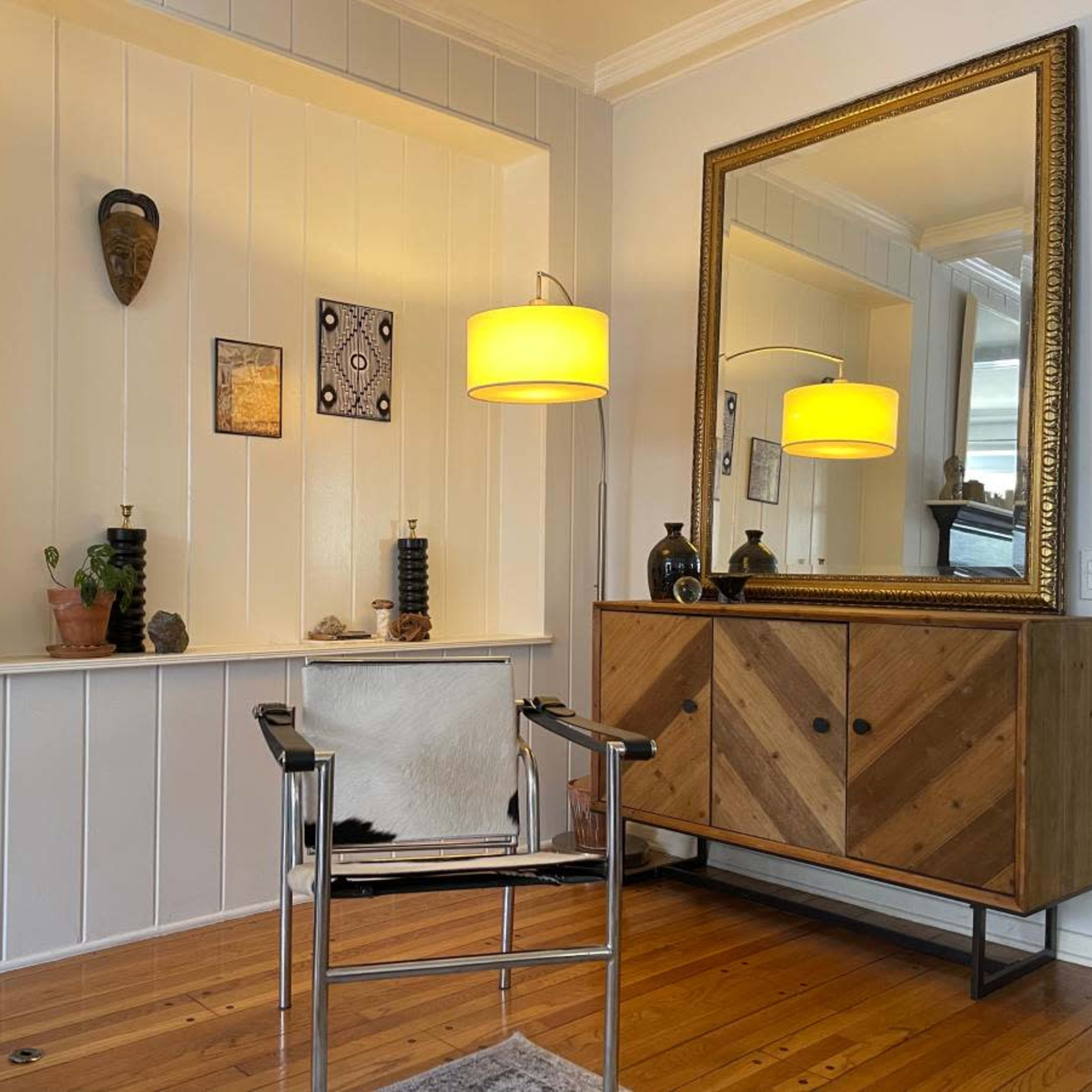 A modern living space features a wooden cabinet, a mirror, a chair, and various decorative items on a shelf against white walls.