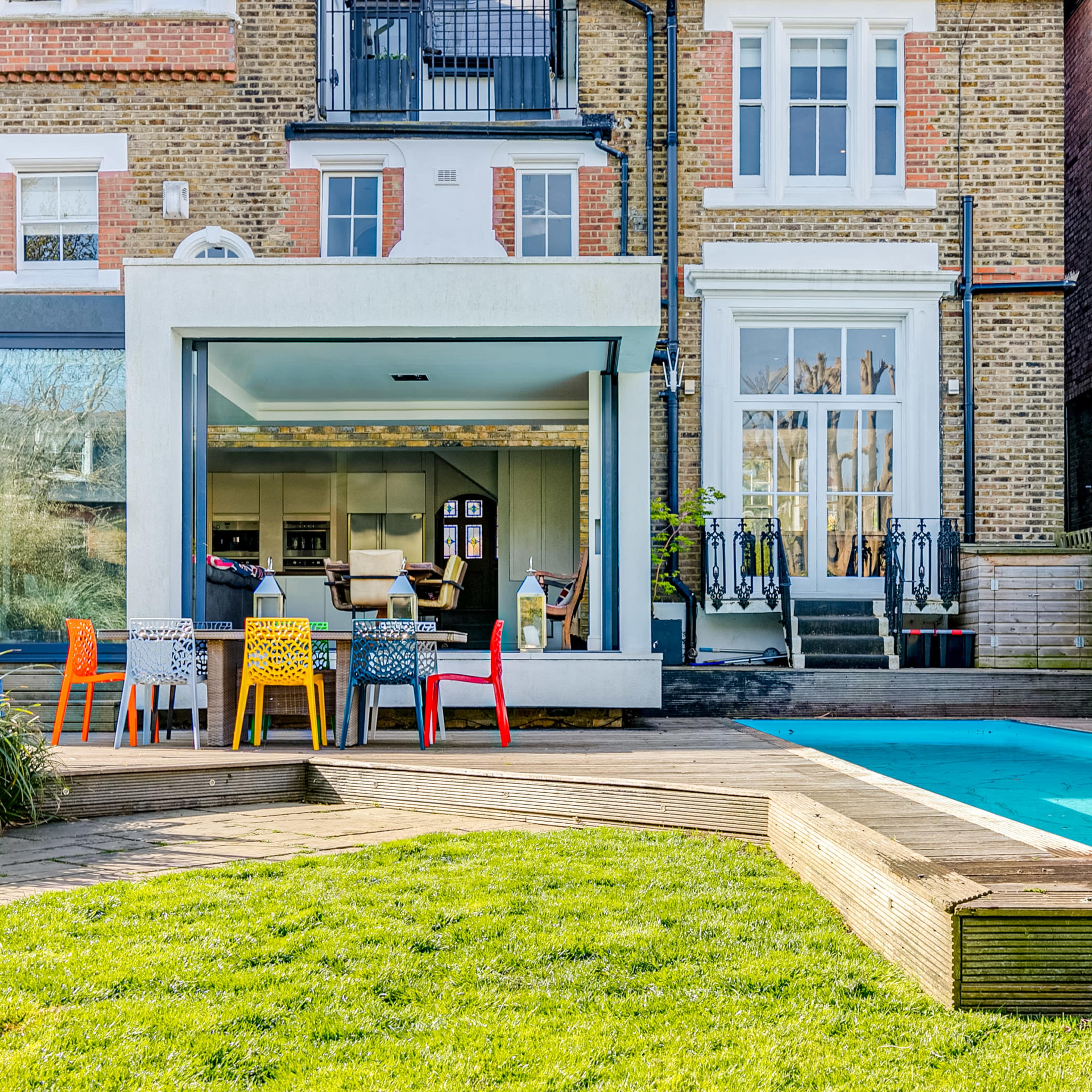 A modern house with a glass extension features a patio area with colorful chairs and a swimming pool in the backyard.