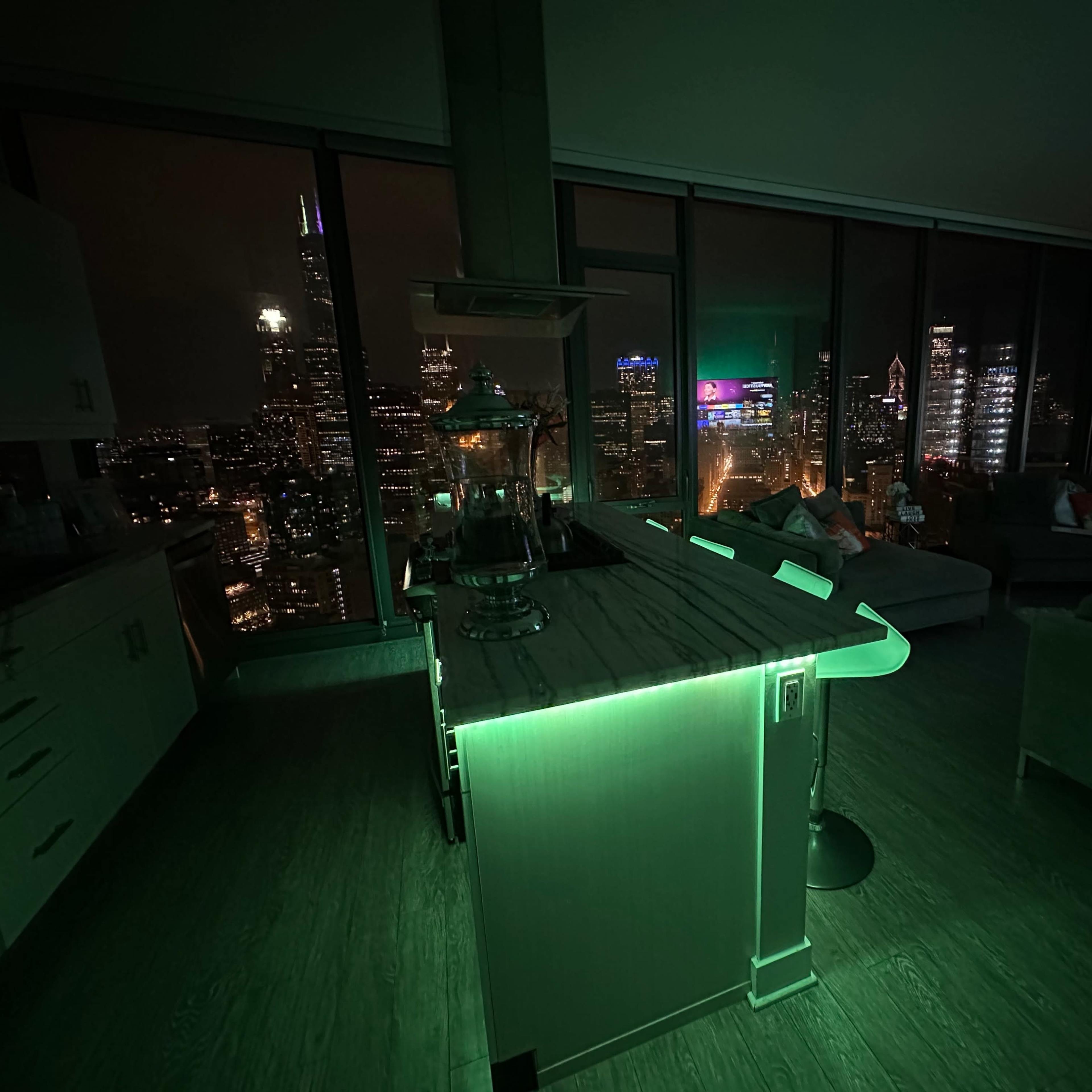 A modern kitchen bar with green LED lighting overlooks a city skyline at night.