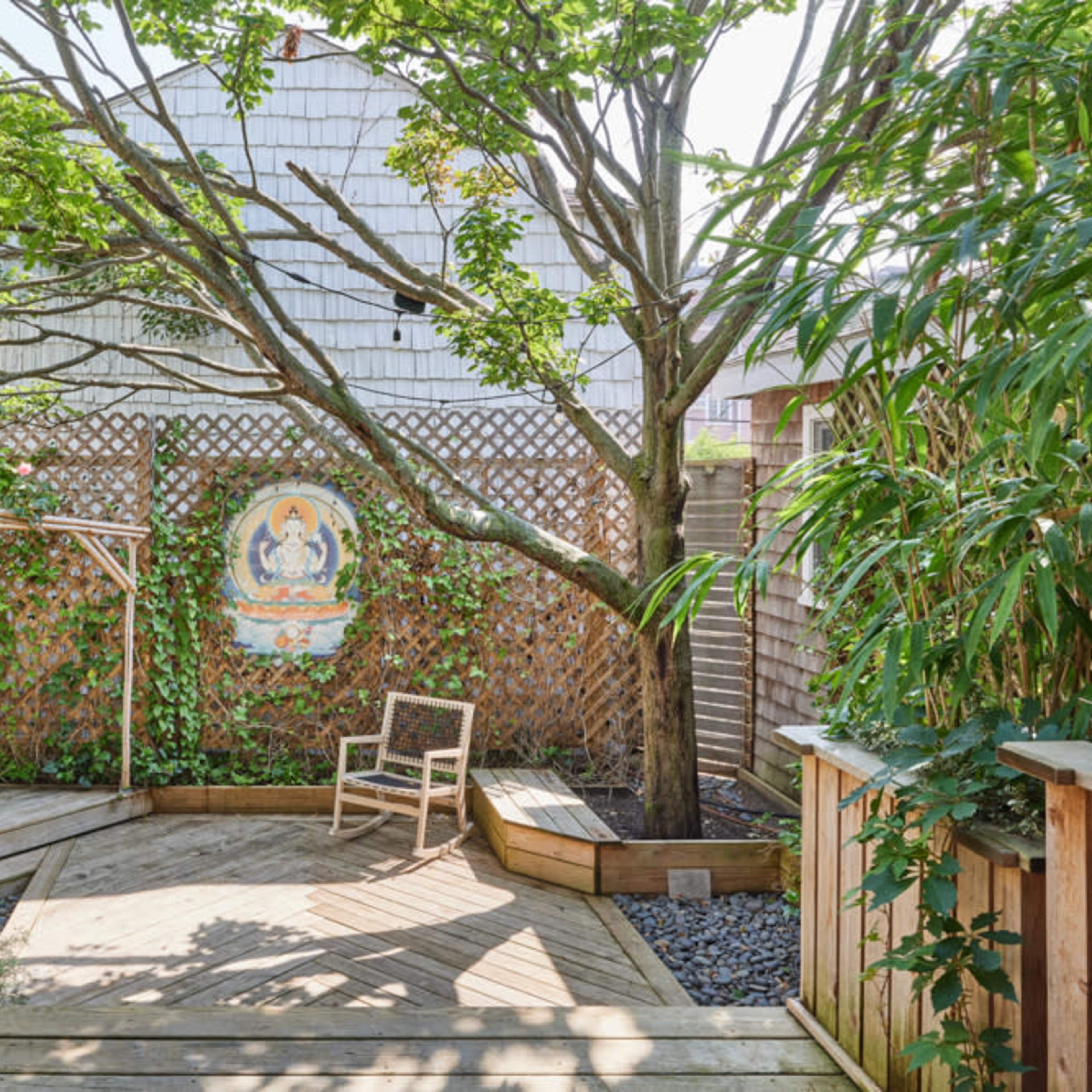 A wooden deck area surrounded by greenery features a single chair and a decorative mural on a lattice wall.