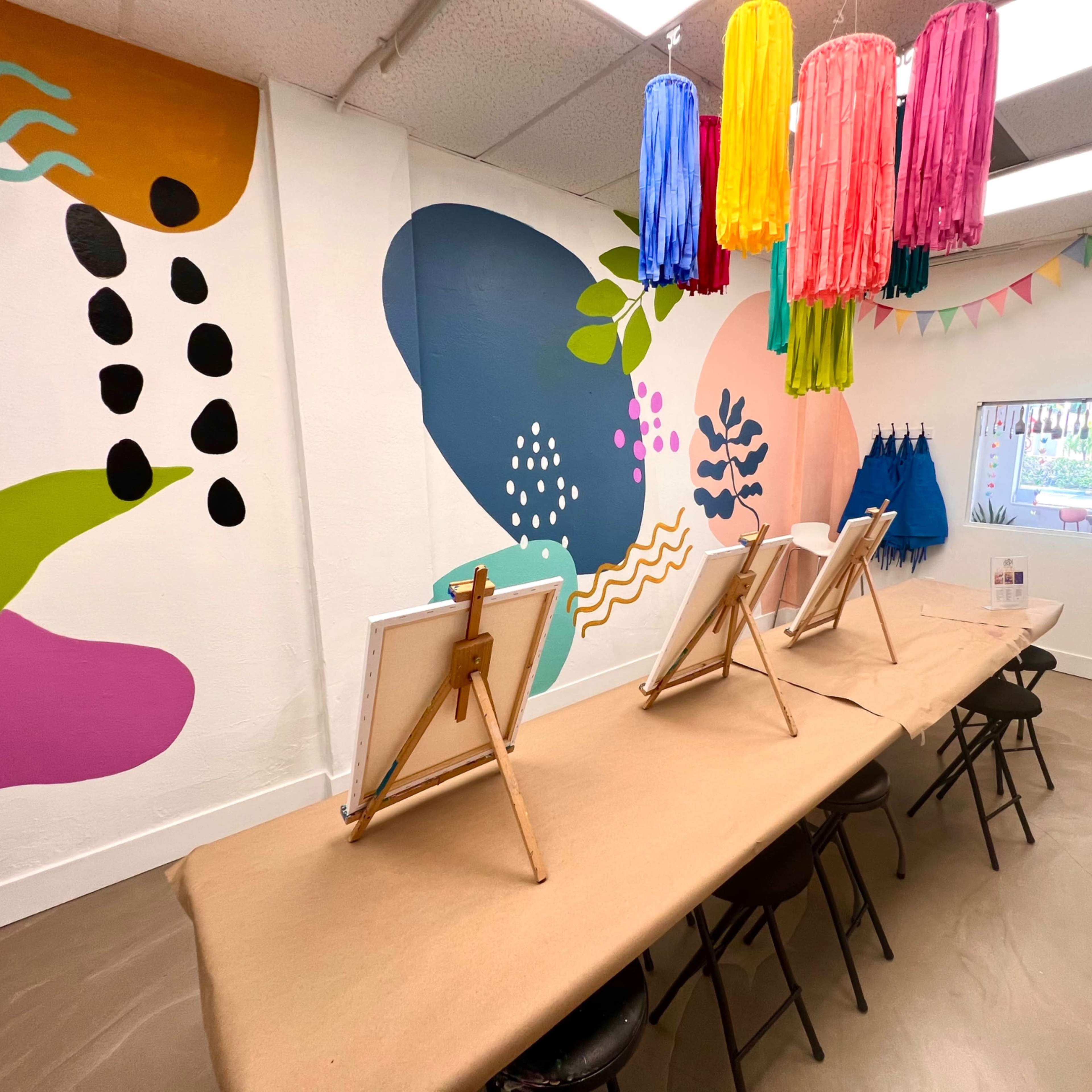 A brightly decorated art studio with a long table set up for painting, featuring easels and colorful paper decorations hanging above.