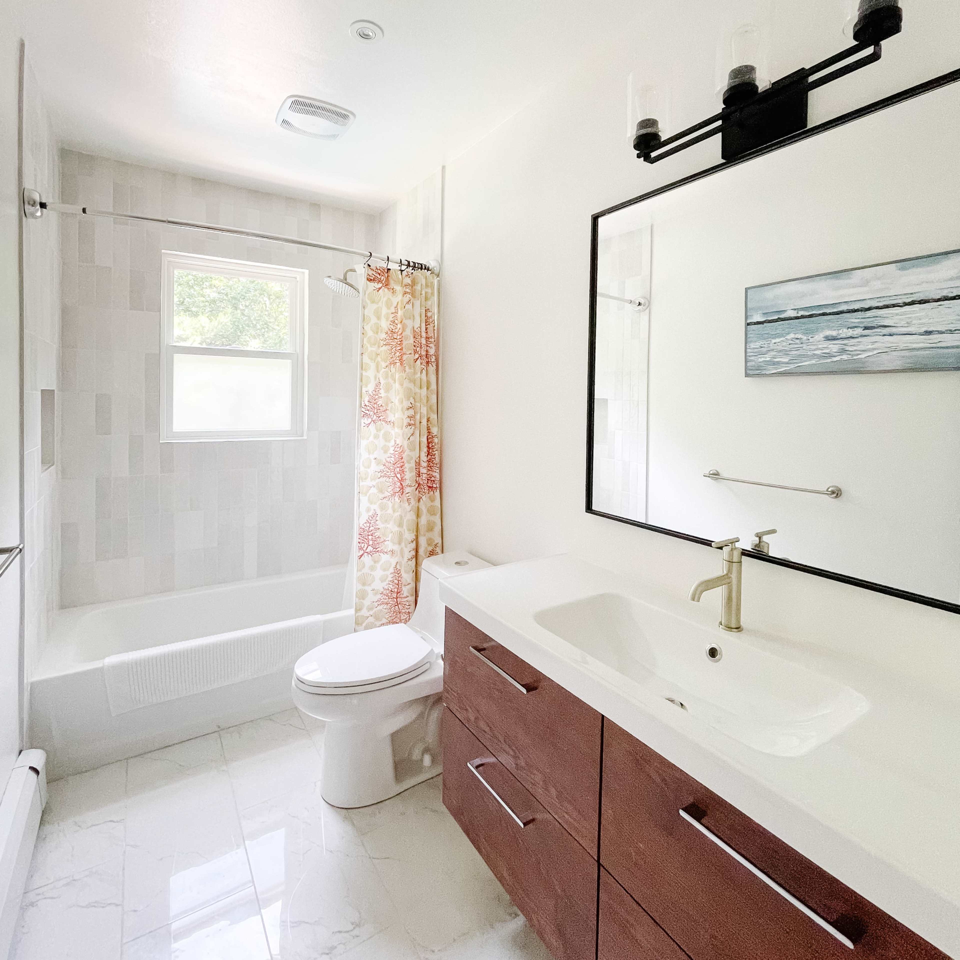 A modern bathroom featuring a bathtub with a shower, a large mirror above a vanity with drawers, and a window providing natural light.