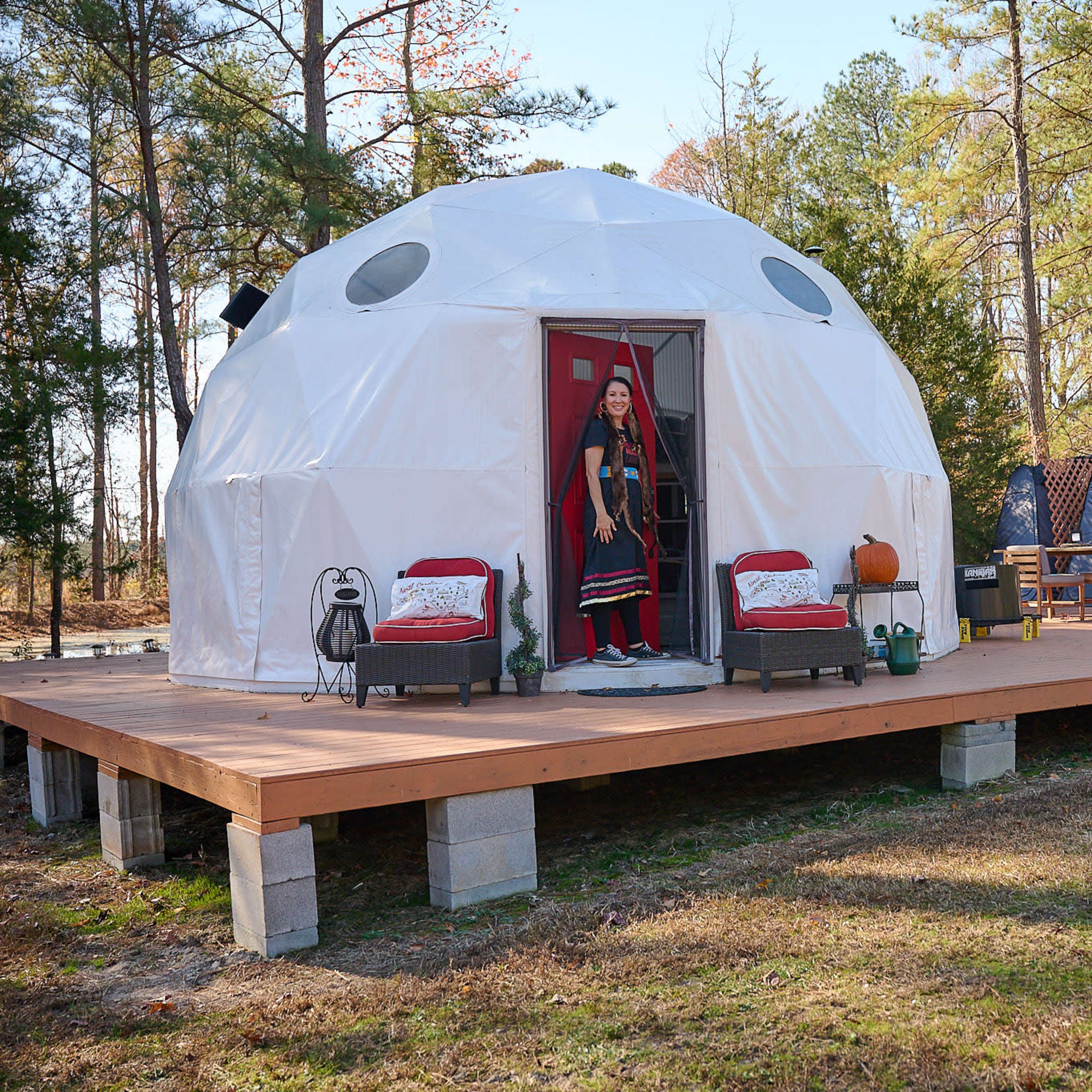 A woman stands in the doorway of a white geodesic dome home situated on a wooden deck surrounded by trees.