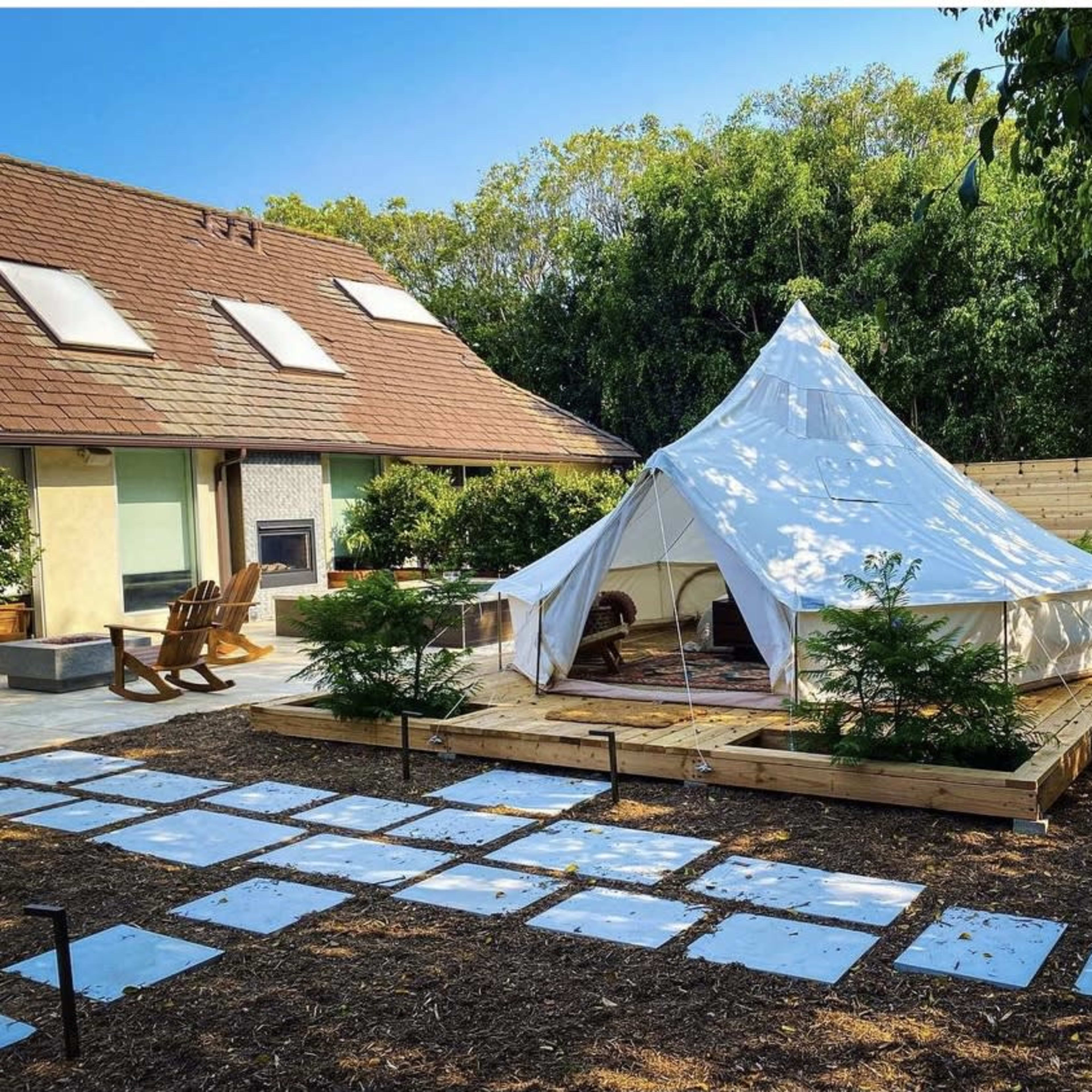 A large white bell tent is set on a wooden platform surrounded by a landscaped area with a stone path leading up to it.