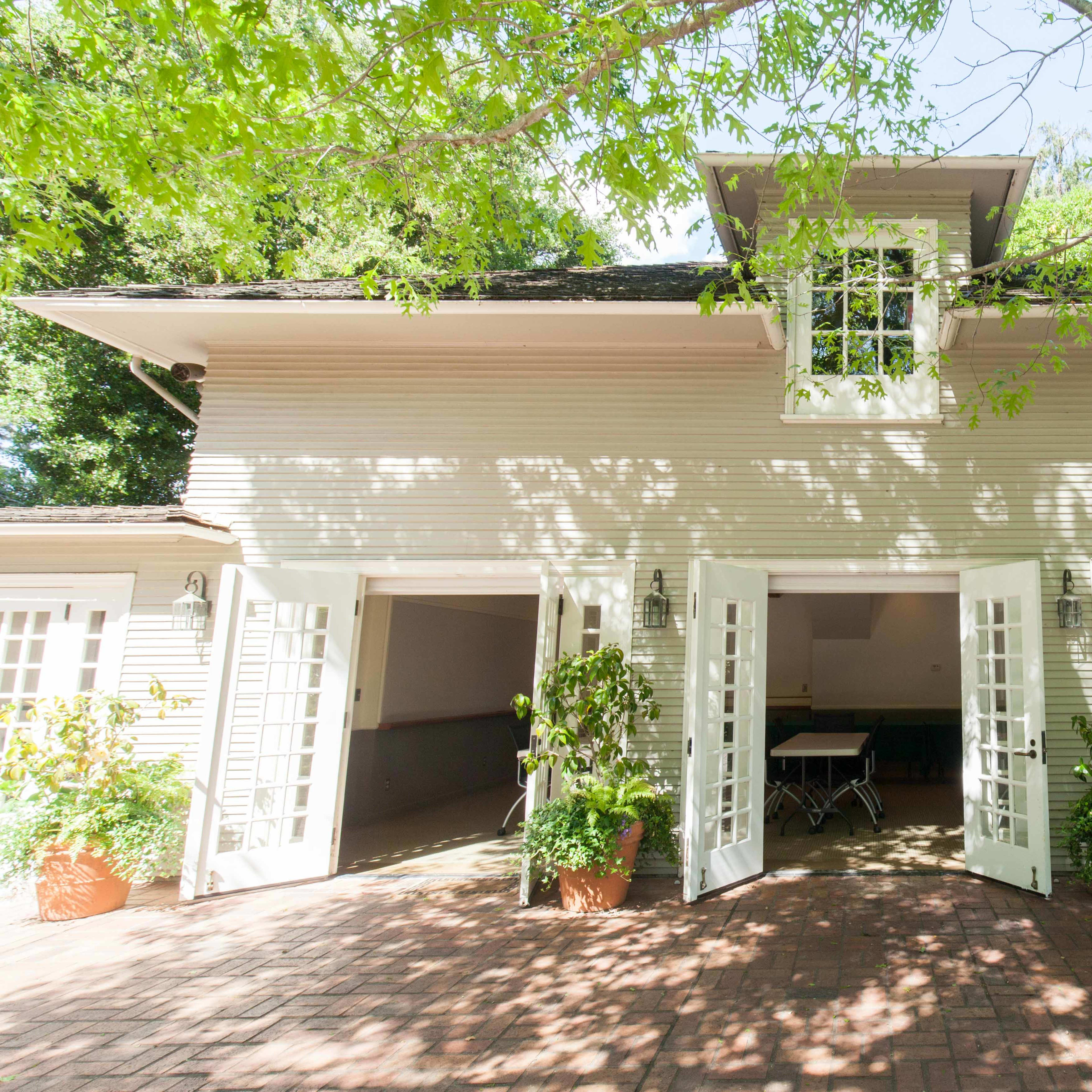 A light-colored building with multiple open French doors and potted plants on a brick patio, framed by leafy trees.
