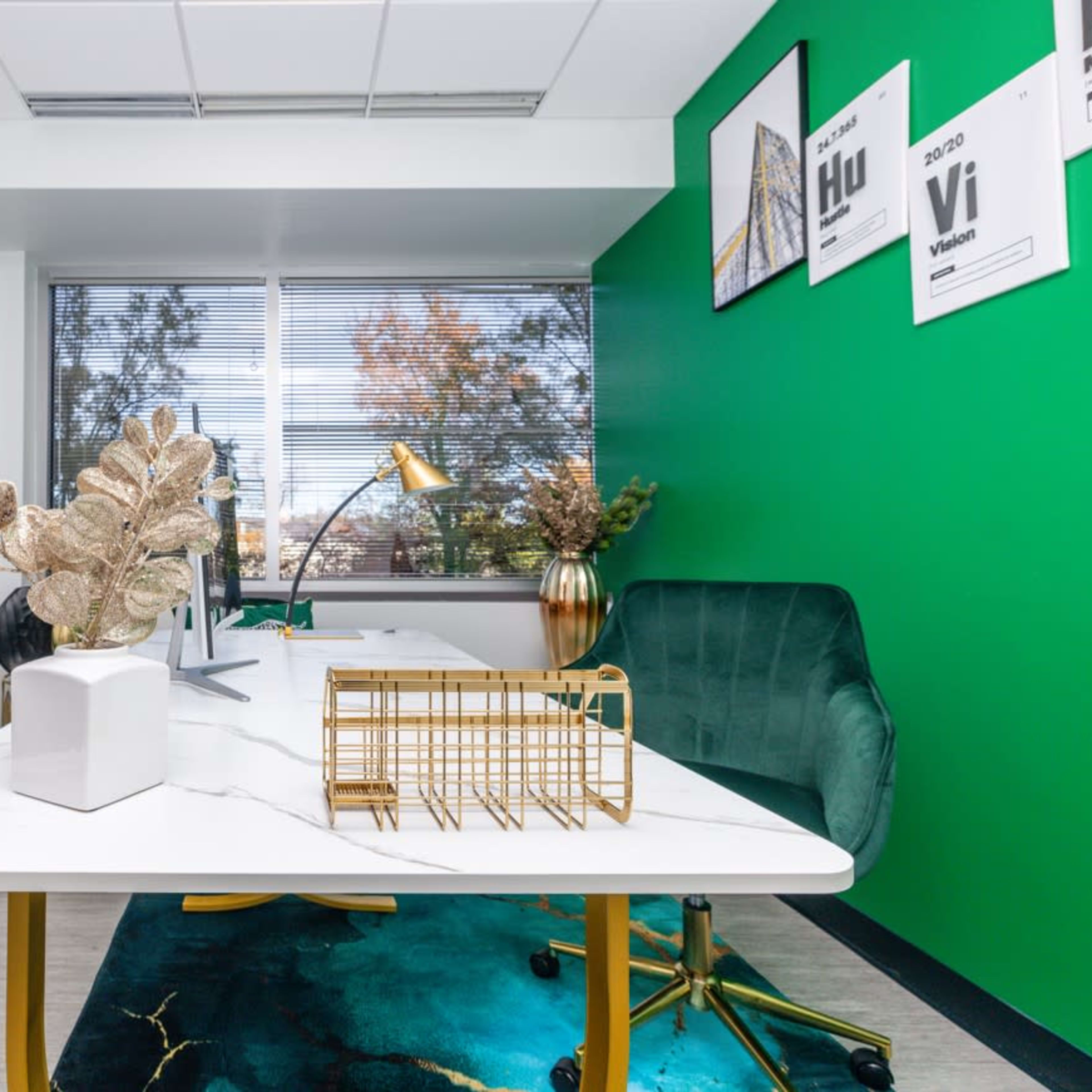 A modern office features a white desk with a gold lamp, green accent walls, and decorative plants, alongside a teal chair and a geometric metal organizer.