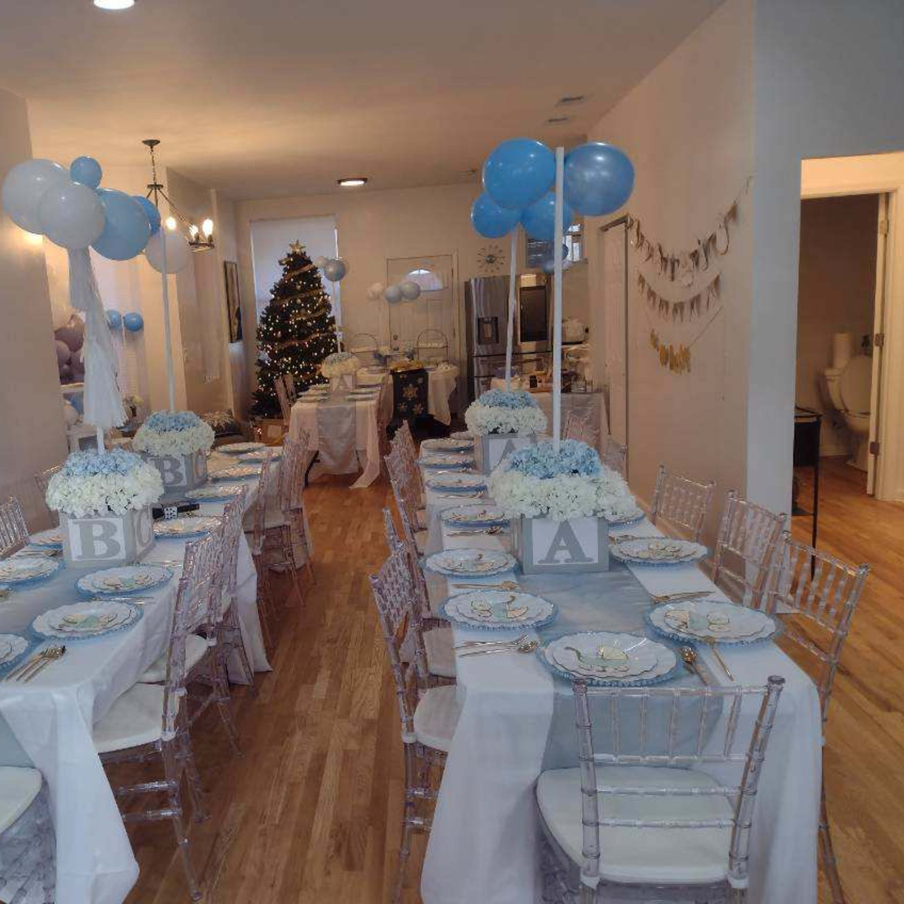 A dining area set up for an event, featuring two long tables decorated with light blue tablecloths, floral centerpieces, and balloons, with a Christmas tree in the background.