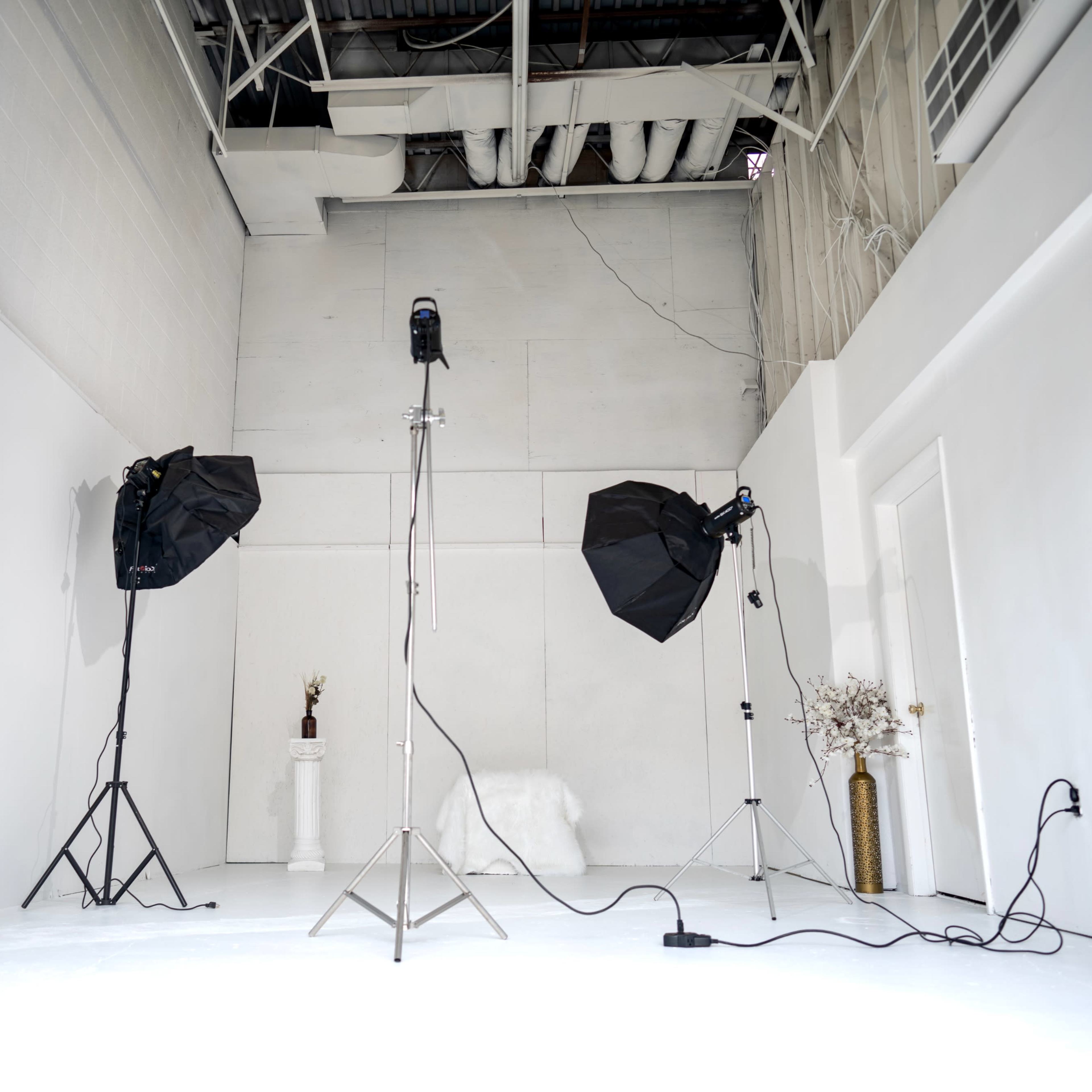 A photography studio with two softbox lights on tripods, a microphone on a stand, and minimal decor, all set against white walls and a white floor.