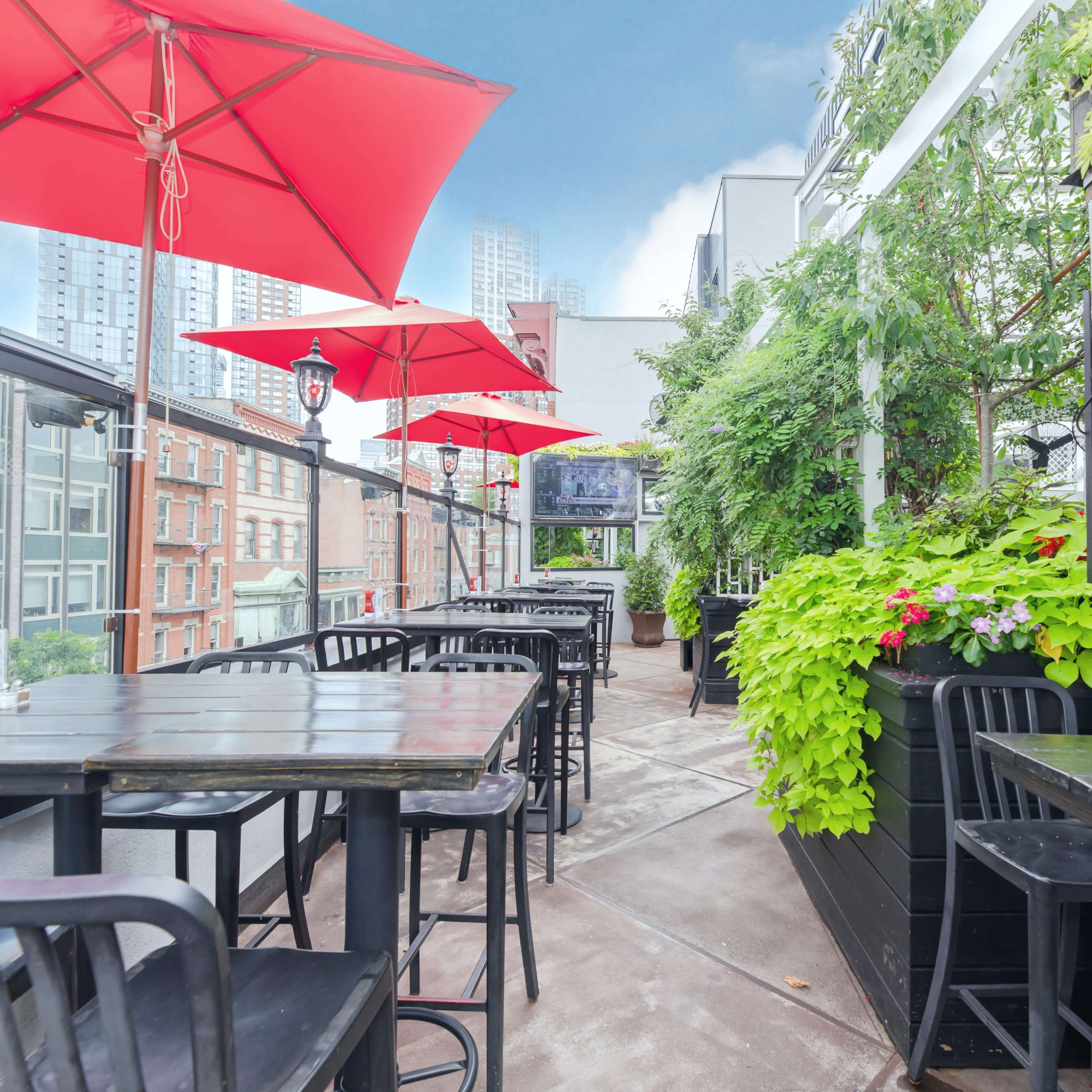 A rooftop terrace features black wooden tables and chairs under red umbrellas, surrounded by greenery and city buildings in the background.
