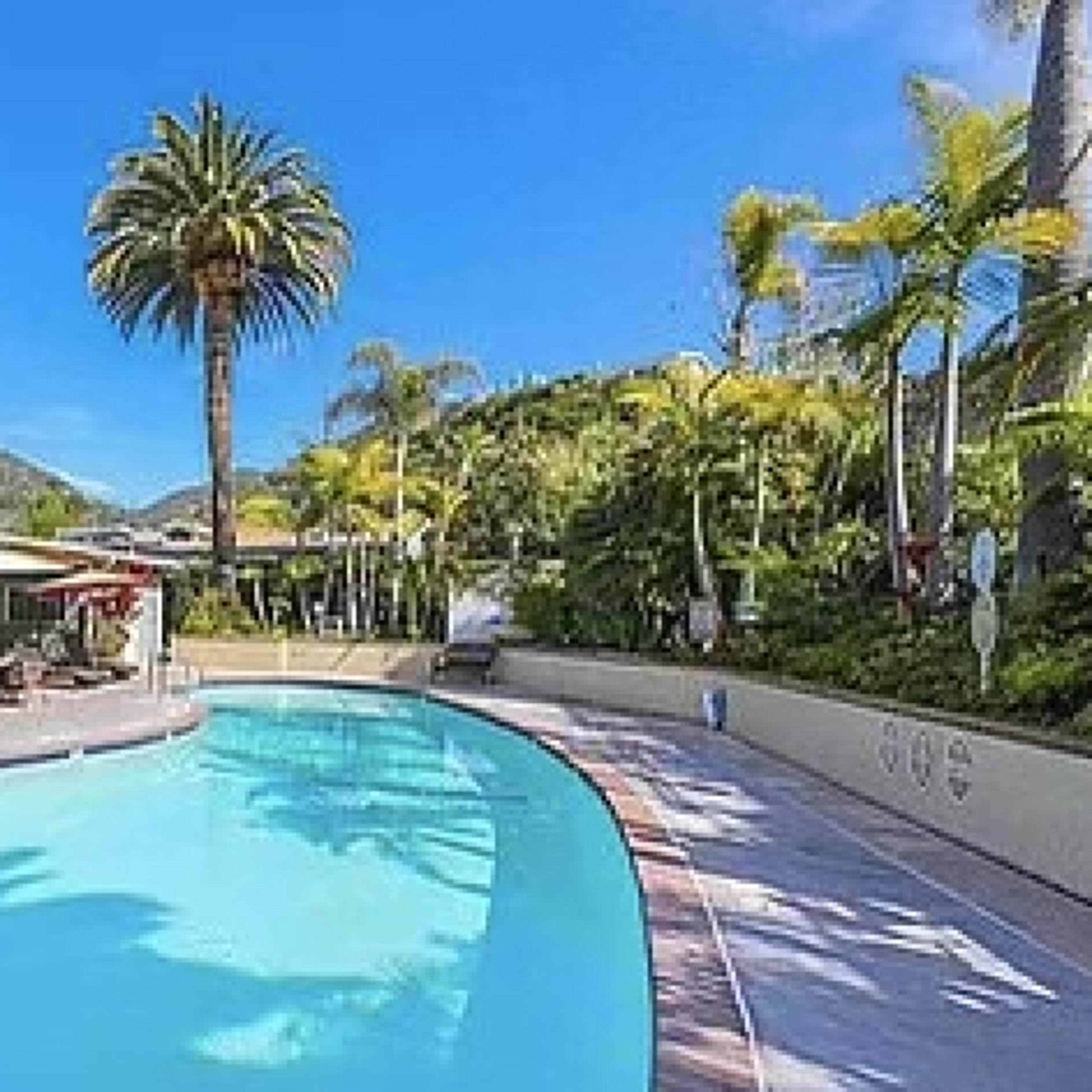 A clear swimming pool is surrounded by palm trees and lounge chairs, with a building featuring shaded tables in the background.