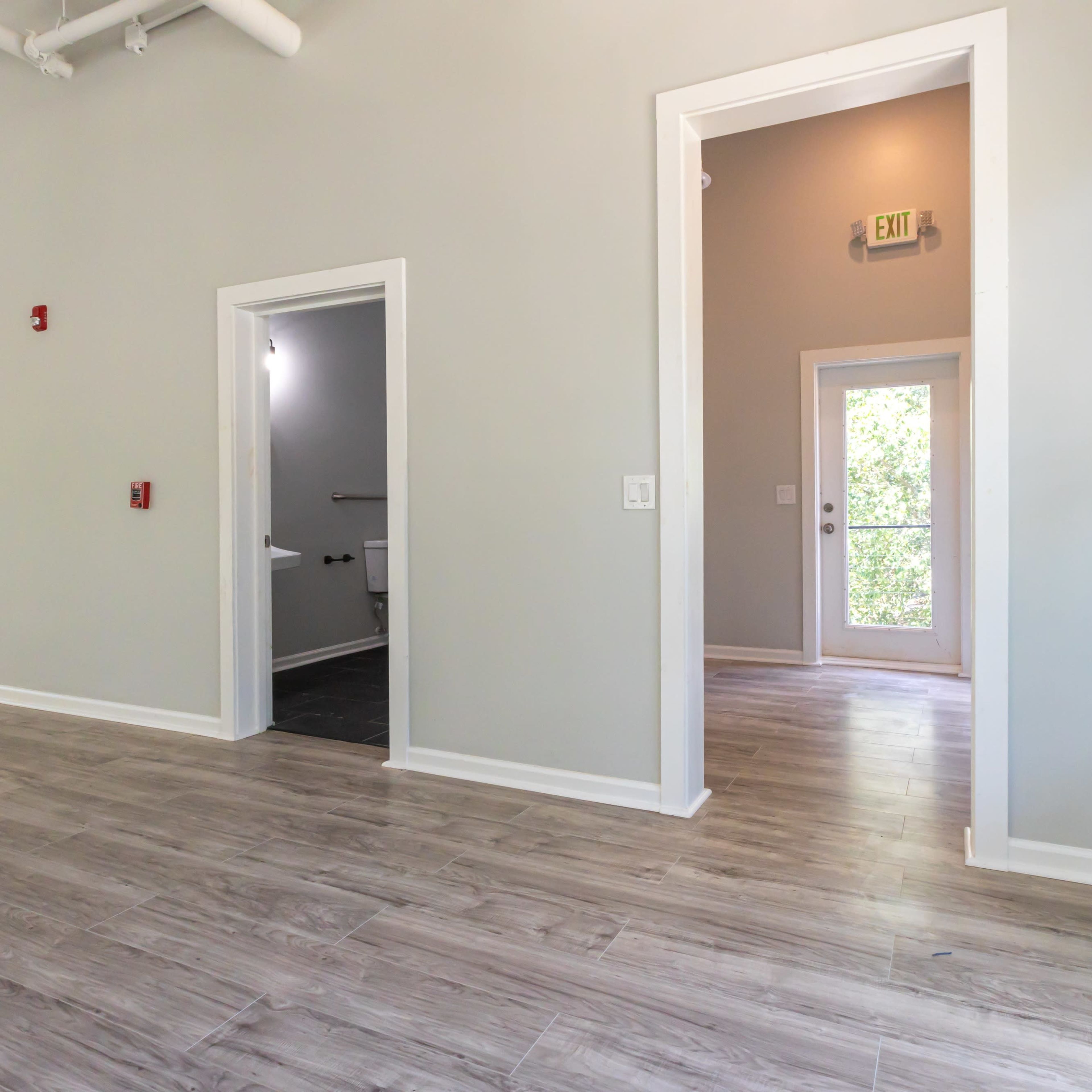A spacious, light-colored room with wooden flooring, featuring doorways leading to other rooms and a view of a door opening to the outdoors.