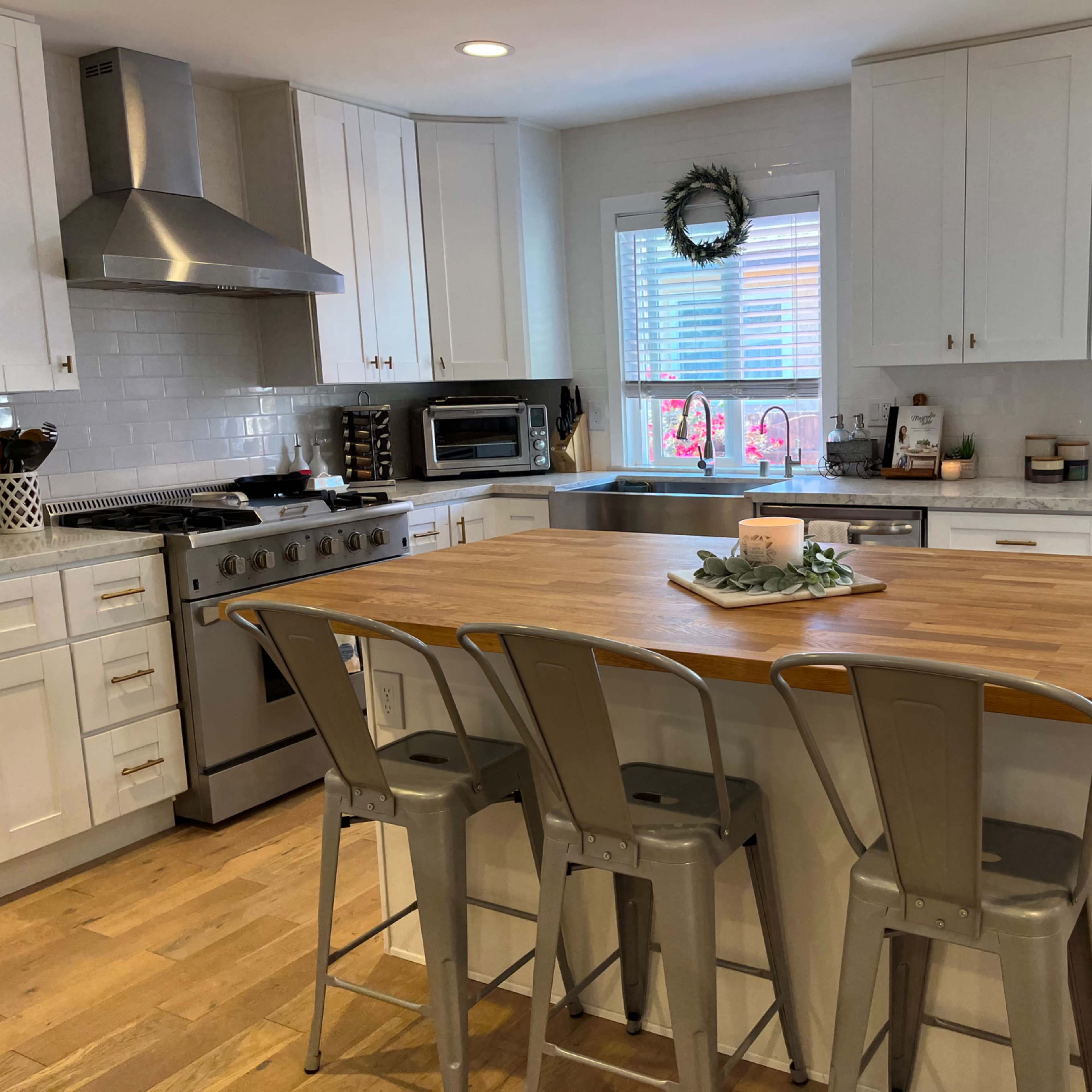 A modern kitchen features white cabinets, a central wooden island with metal stools, and a window overlooking a garden.