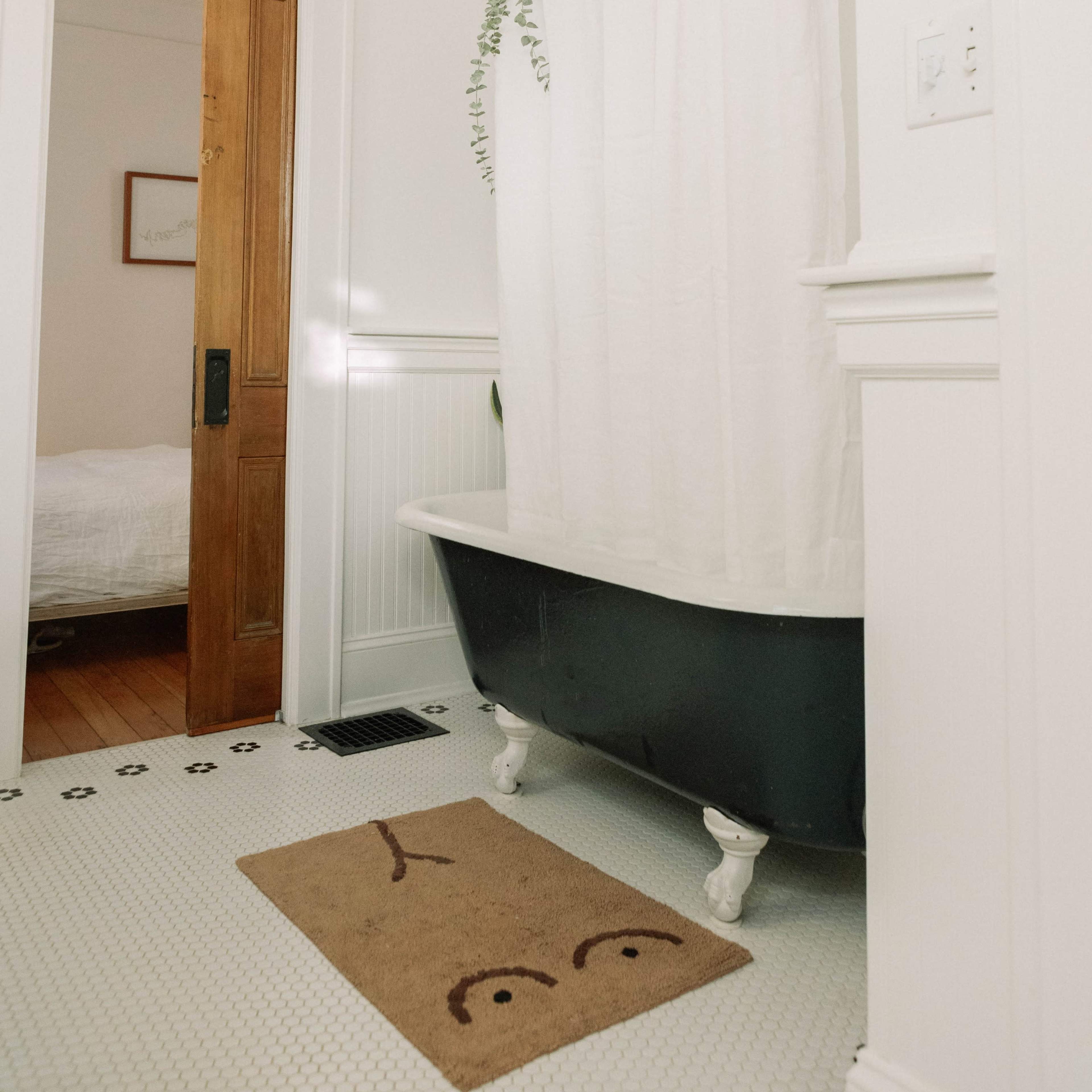 A bathroom featuring a black clawfoot bathtub with a white shower curtain, a welcome mat on the floor, and a wooden door leading to another room.