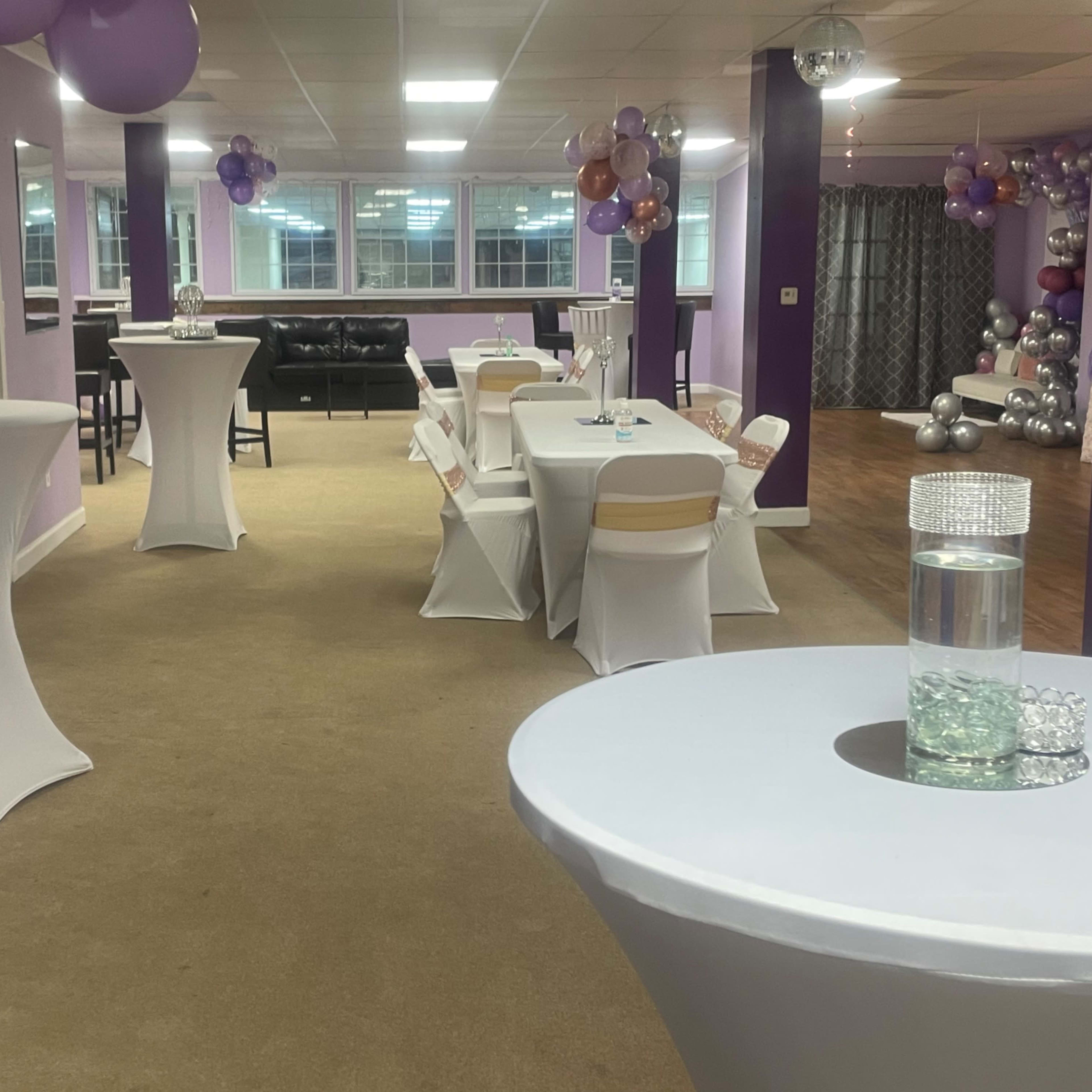 A party venue with tables dressed in white covers, purple and silver decorations, and a spacious area filled with balloons and a mirrored backdrop.