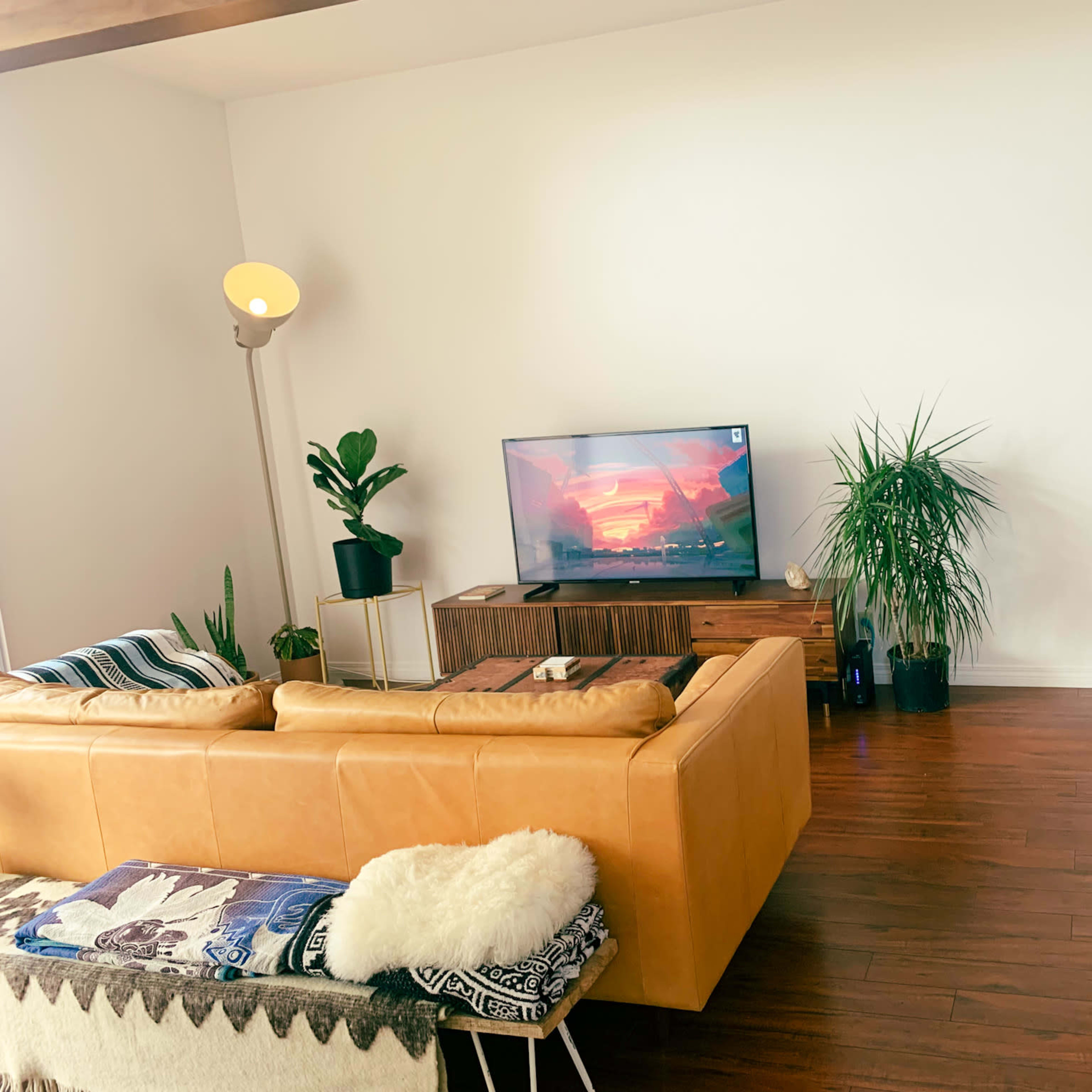 A cozy living room features a beige leather couch, a wooden TV stand with a television displaying a colorful scene, and potted plants alongside a doorway leading to another room.