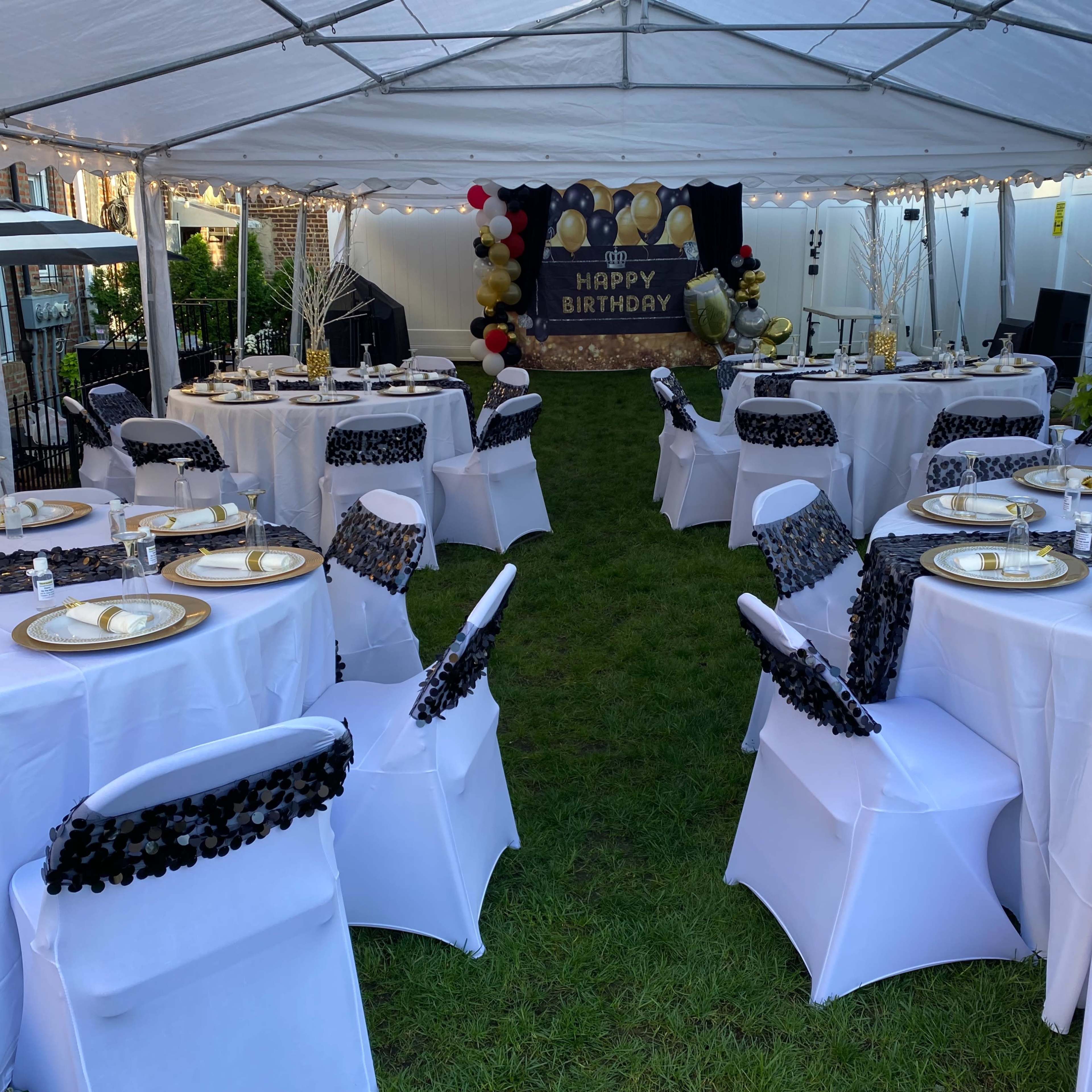 A tented outdoor space features tables set with white tablecloths and gold plates, adorned with black trim, alongside a "Happy Birthday" backdrop.