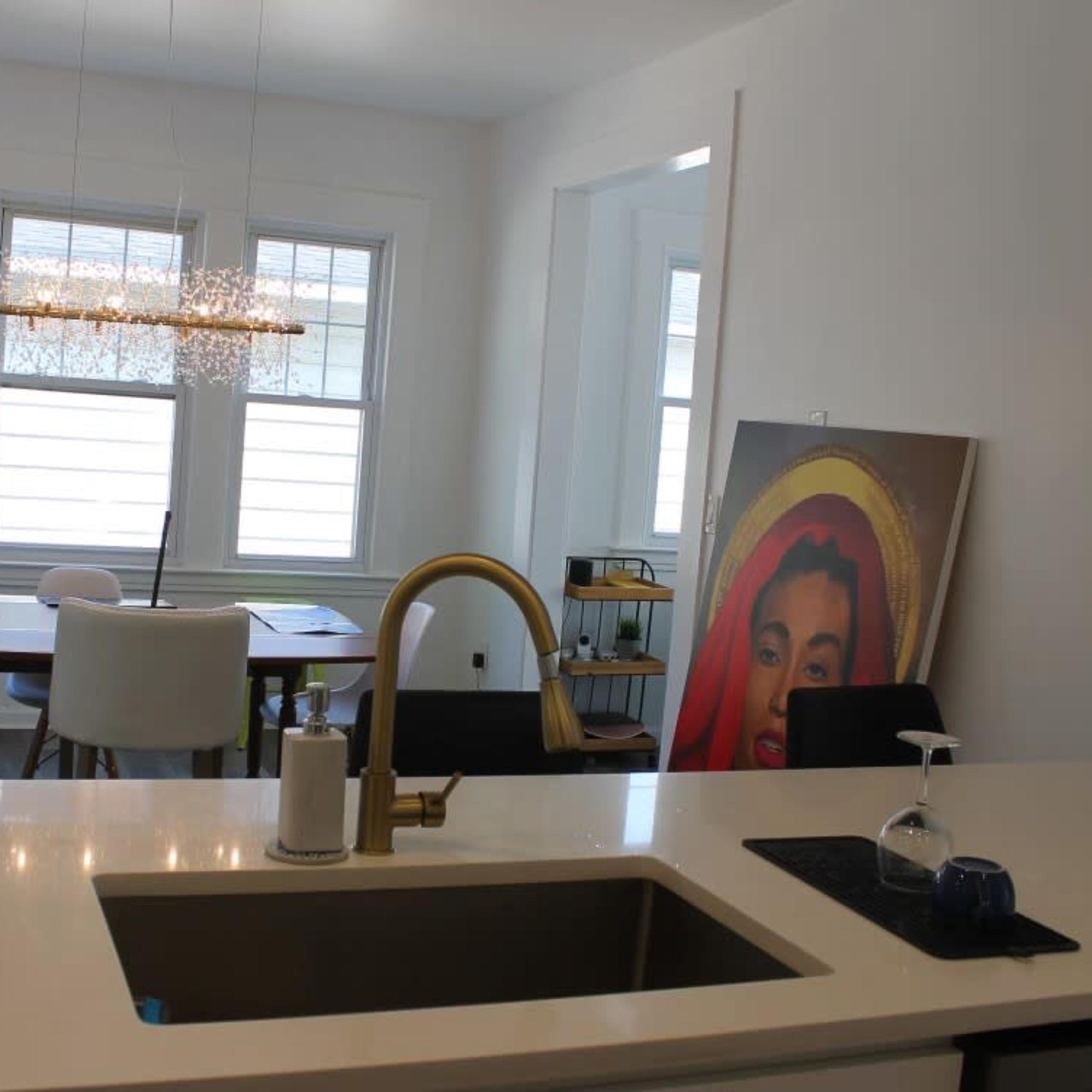 A modern kitchen with a sink, leading into a dining area with a table and chairs, illuminated by natural light from windows.