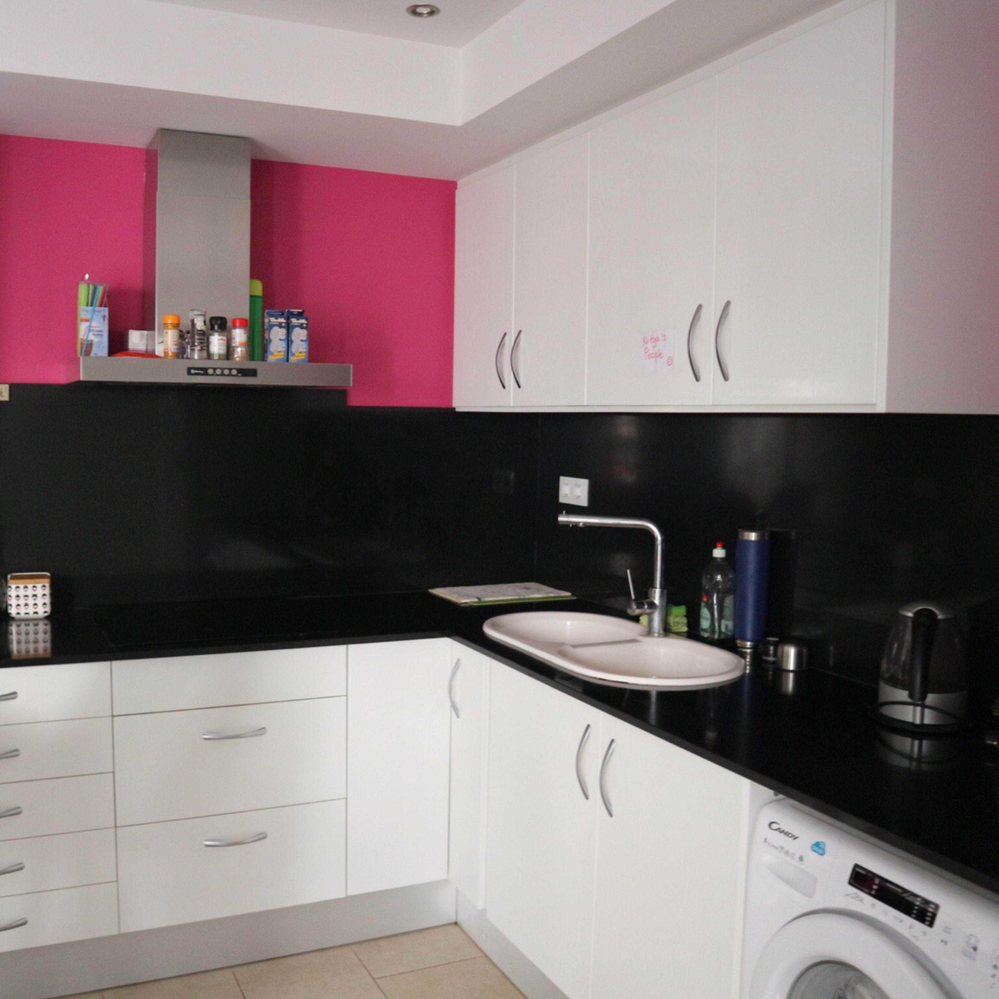 A modern kitchen features black countertops, white cabinetry, a pink accent wall, and stainless steel appliances.