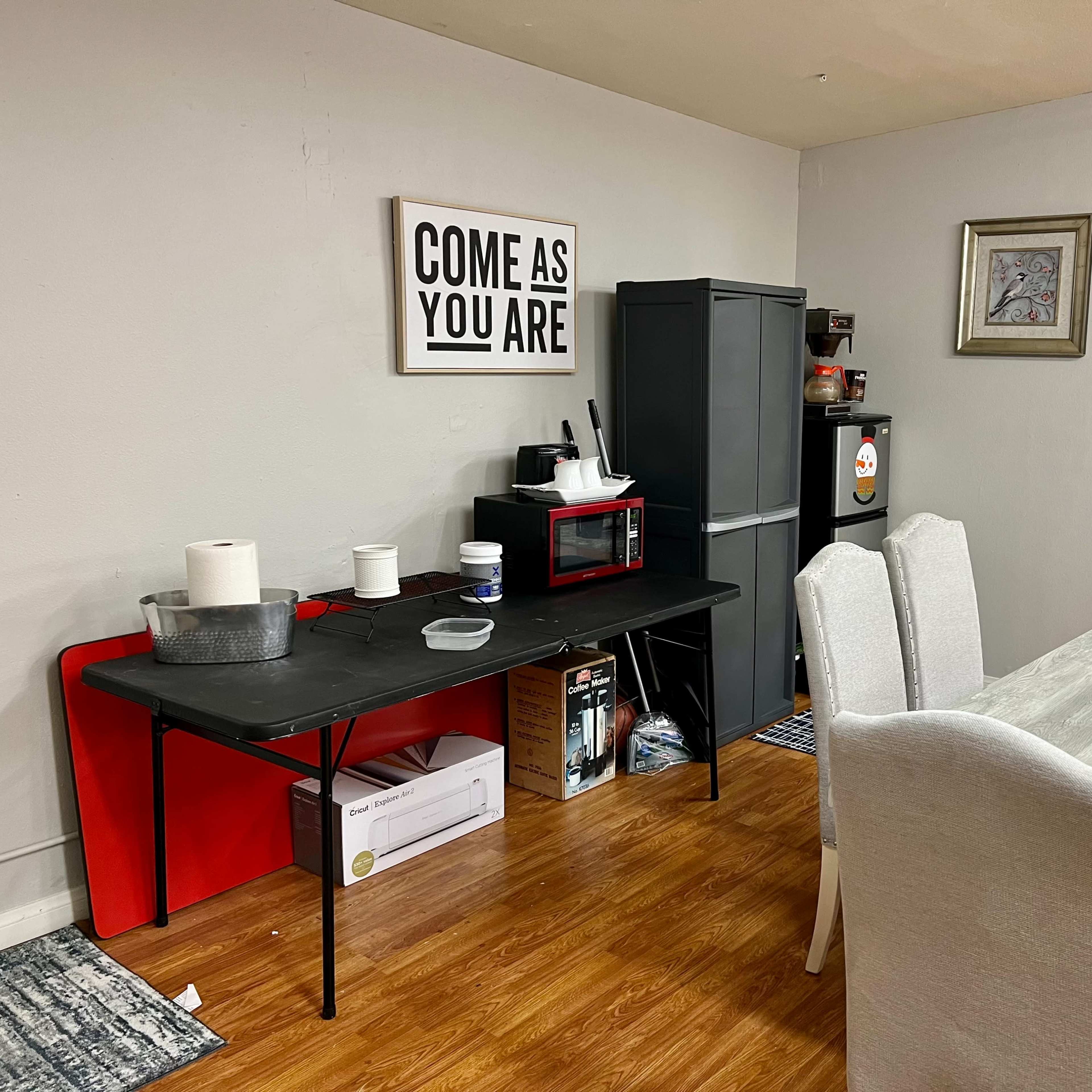A dining area with a gray table and chairs, a black table with kitchen items, and a small kitchen appliance setup against a wall featuring a large framed sign that reads "COME AS YOU ARE."