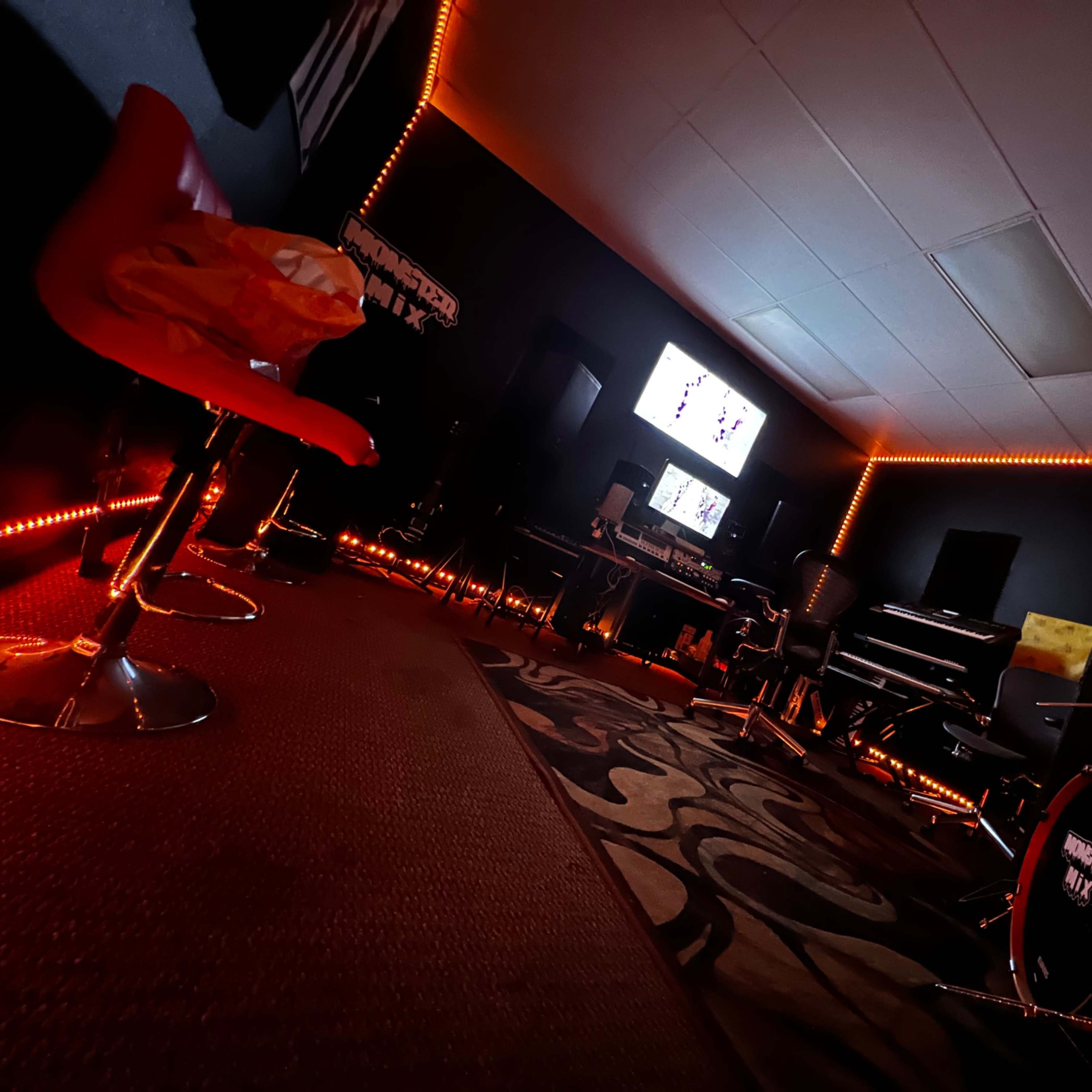 A music studio featuring a drum set, a piano, a computer setup, and red accent lighting along the walls.