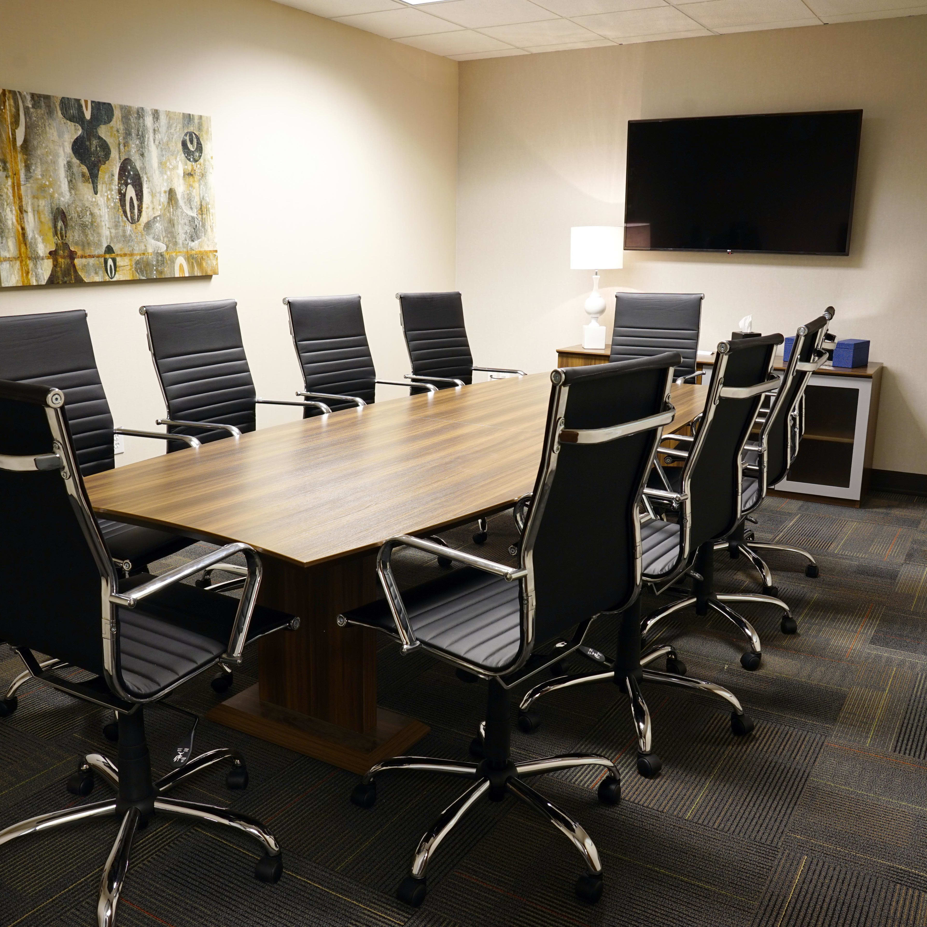 A conference room features a large wooden table surrounded by black office chairs, with a television mounted on the wall and artwork displayed.