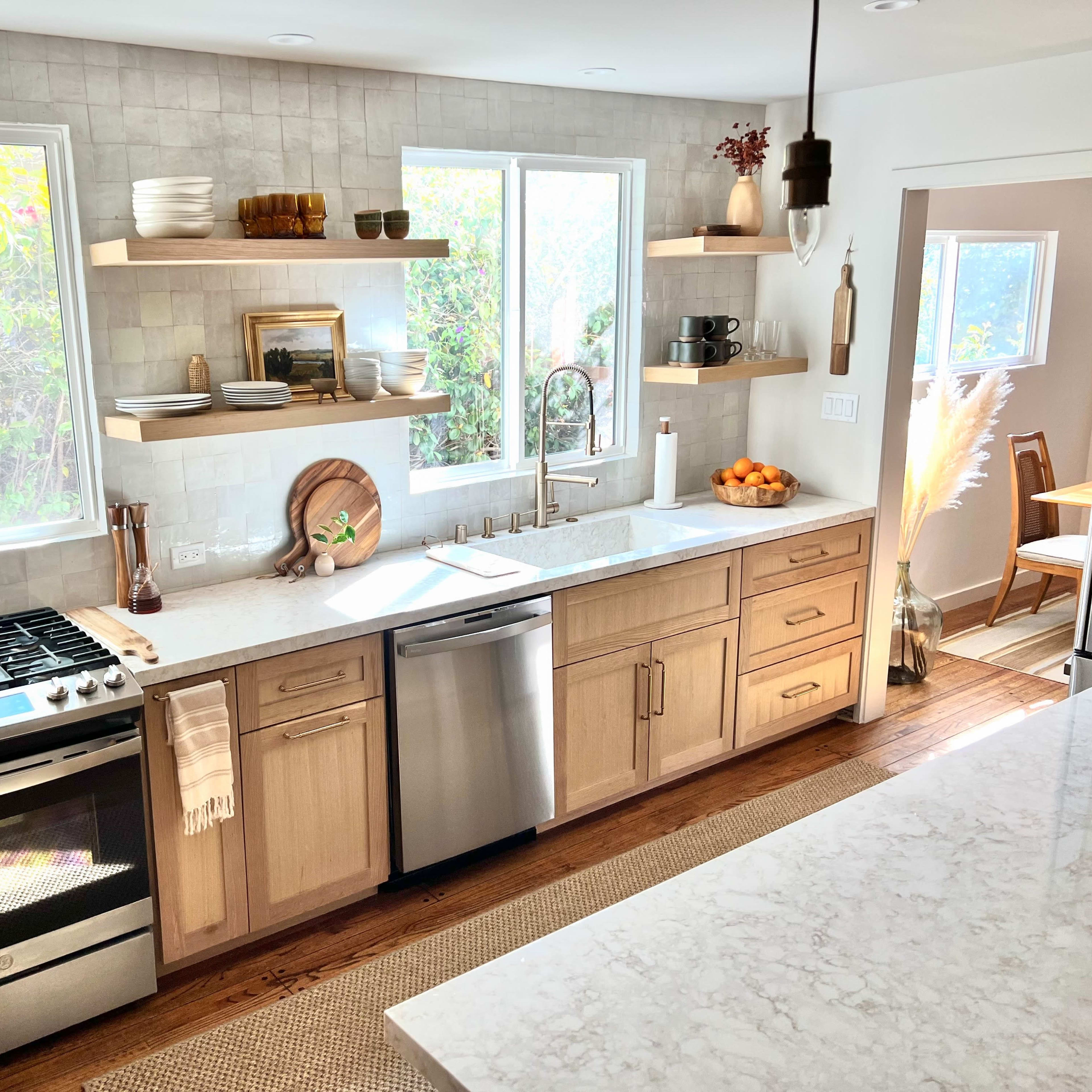 A bright kitchen features wooden shelves with decorative items, a stainless steel stove, and a large marble countertop with a bowl of oranges.