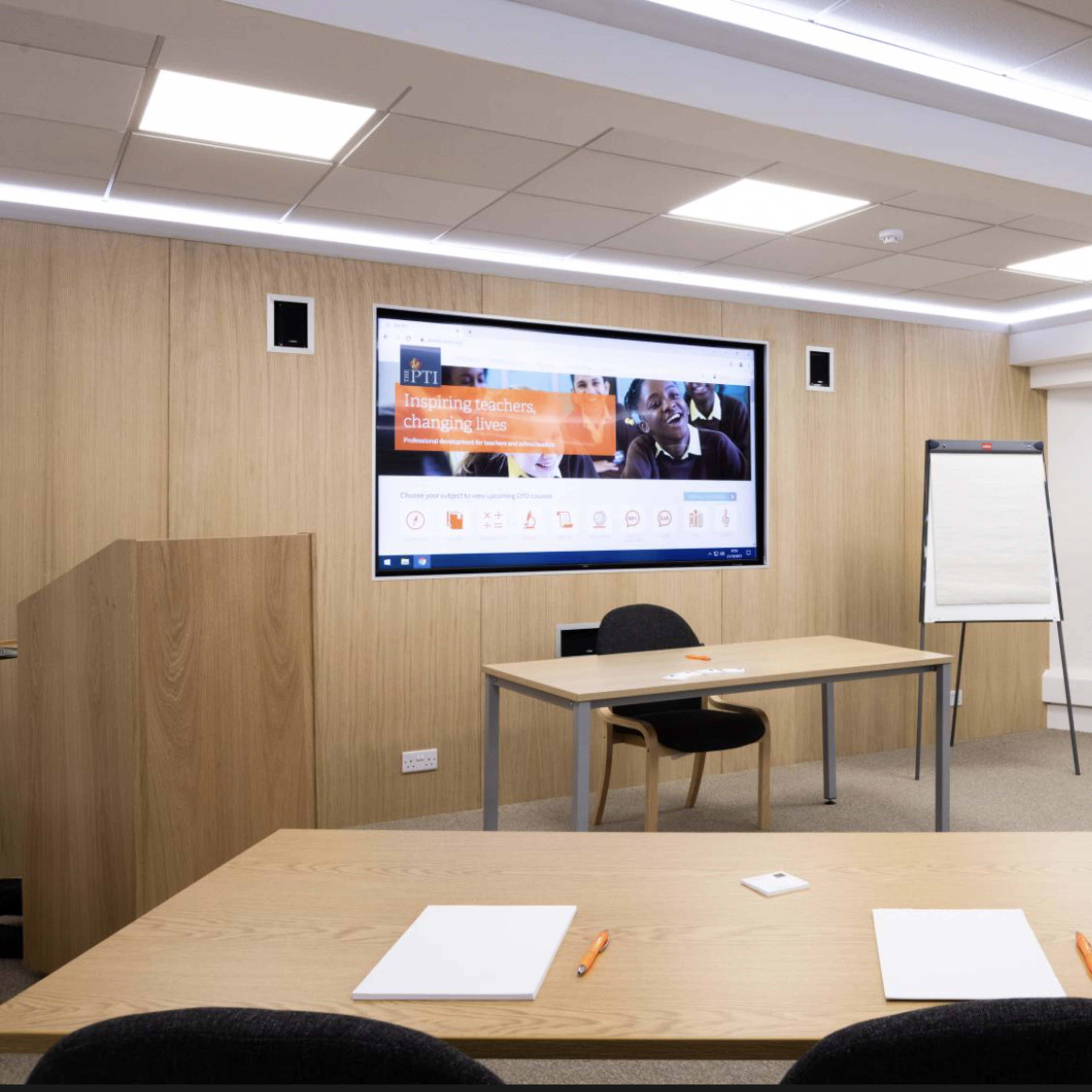 A modern meeting room featuring a large screen, a wooden presentation podium, a table with chairs, and a whiteboard.