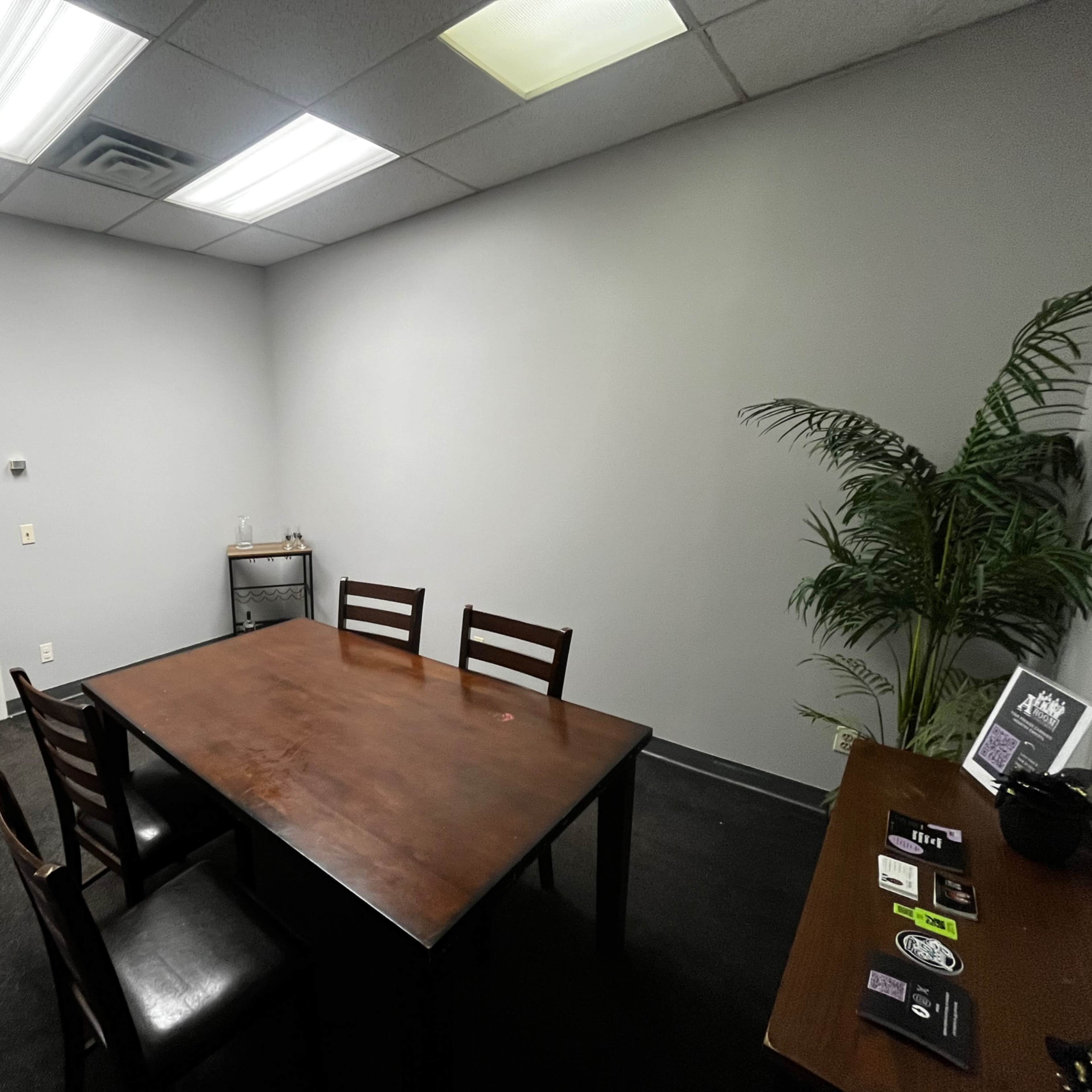 A small meeting room features a brown table surrounded by four chairs, a decorative plant in the corner, and a brown door leading outside.